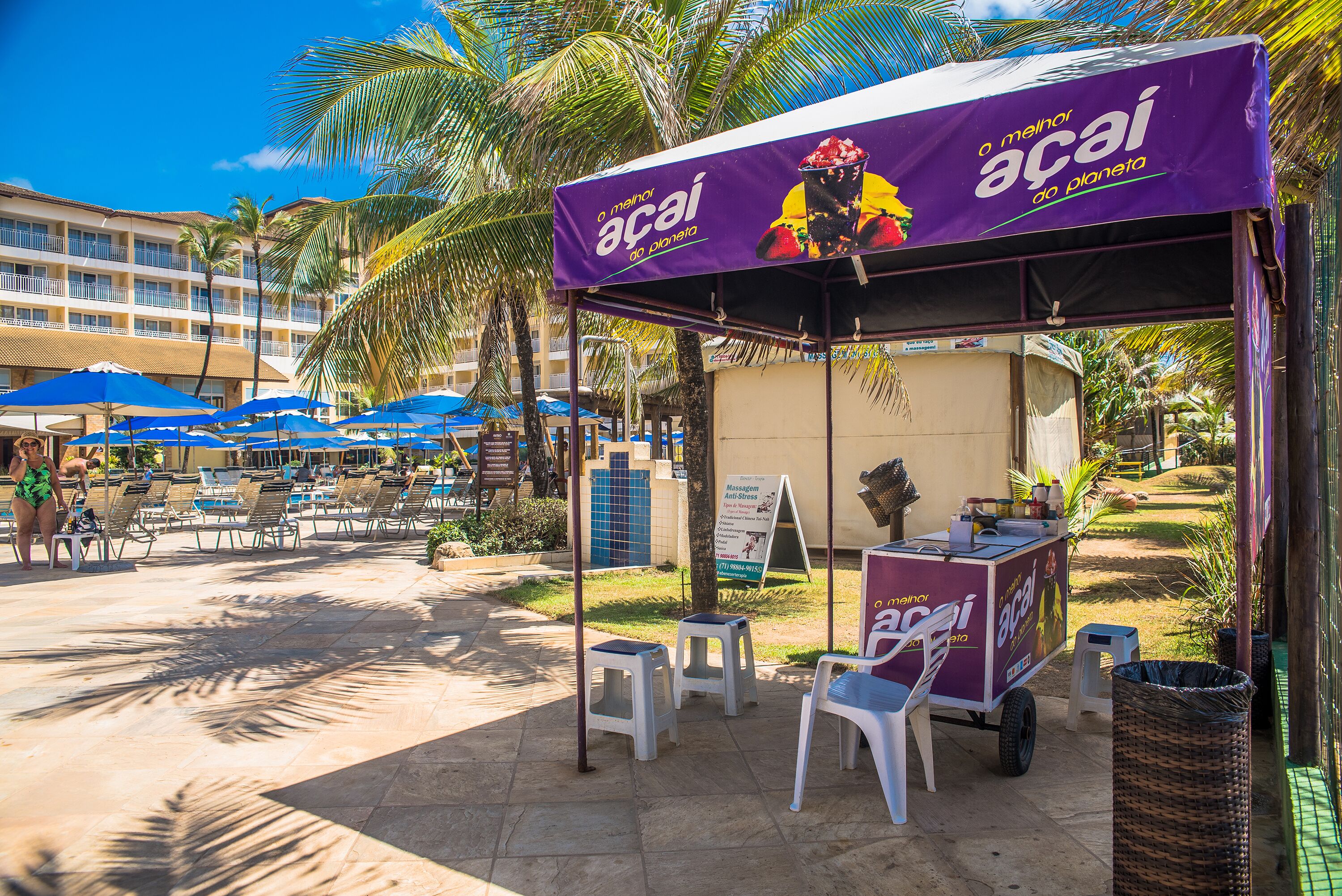 Un carrito de comida en la playa con un toldo morado que anuncia 'Acaí', rodeado de sillones de descanso y palmeras, ofreciendo bebidas de frutas tropicales.
