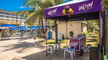 Un carrito de comida en la playa con un toldo morado que anuncia 'Acaí', rodeado de sillones de descanso y palmeras, ofreciendo bebidas de frutas tropicales.