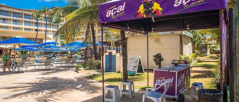A beachside food cart with a purple canopy advertising 'Acaí' with tropical fruit drinks, surrounded by lounge chairs and palm trees.
