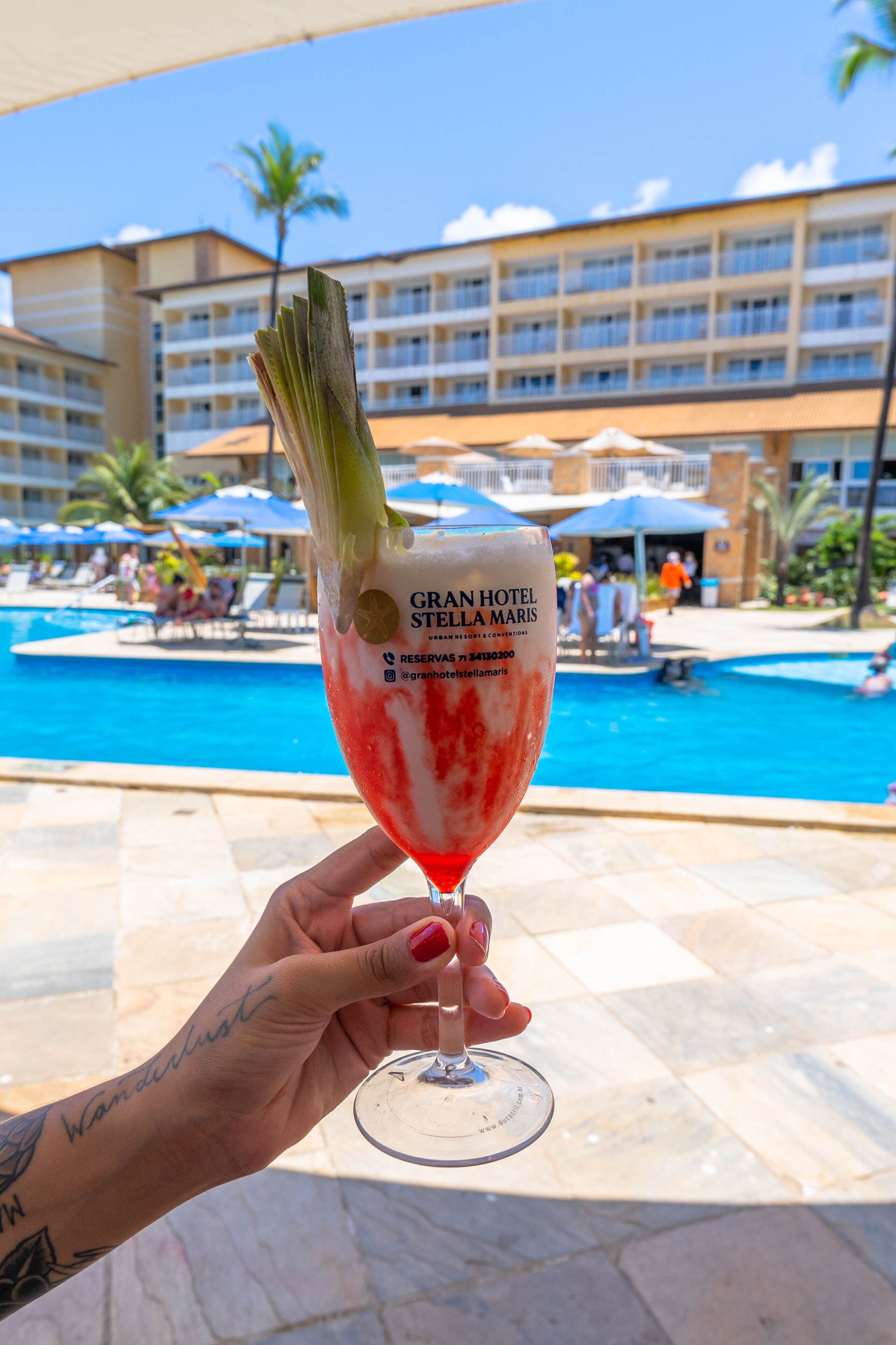 Una mano sosteniendo una copa de cóctel con adorno, frente a un área de piscina de hotel con vista al edificio Gran Hotel Stella Maris.