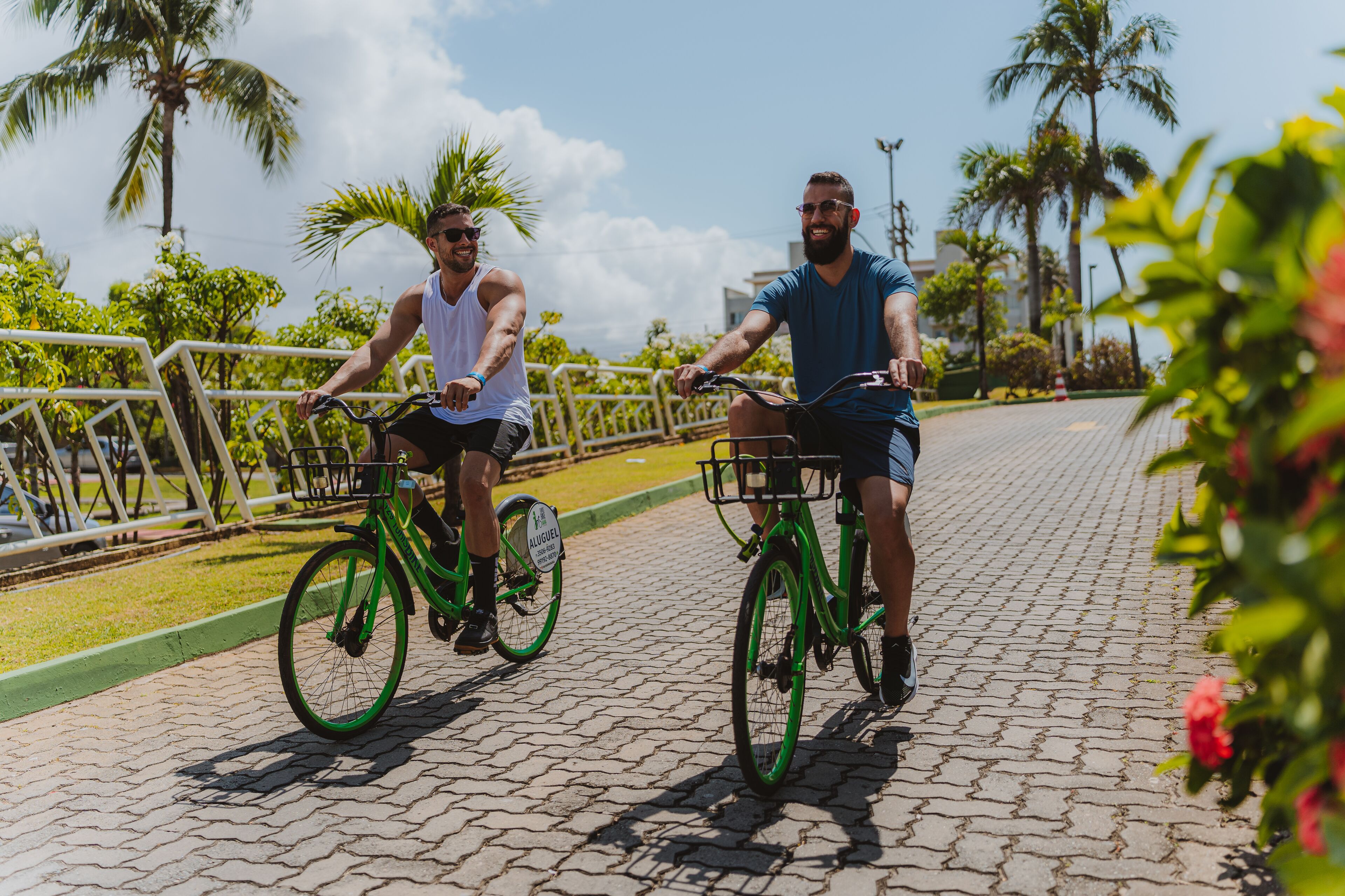 Dos personas andando en bicicleta por un camino pavimentado, con palmeras y follaje al fondo.