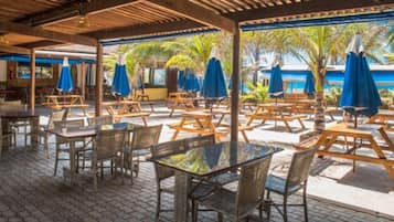Um café ao ar livre com mesas e cadeiras de madeira, guarda-sóis azuis e piso de azulejos.
