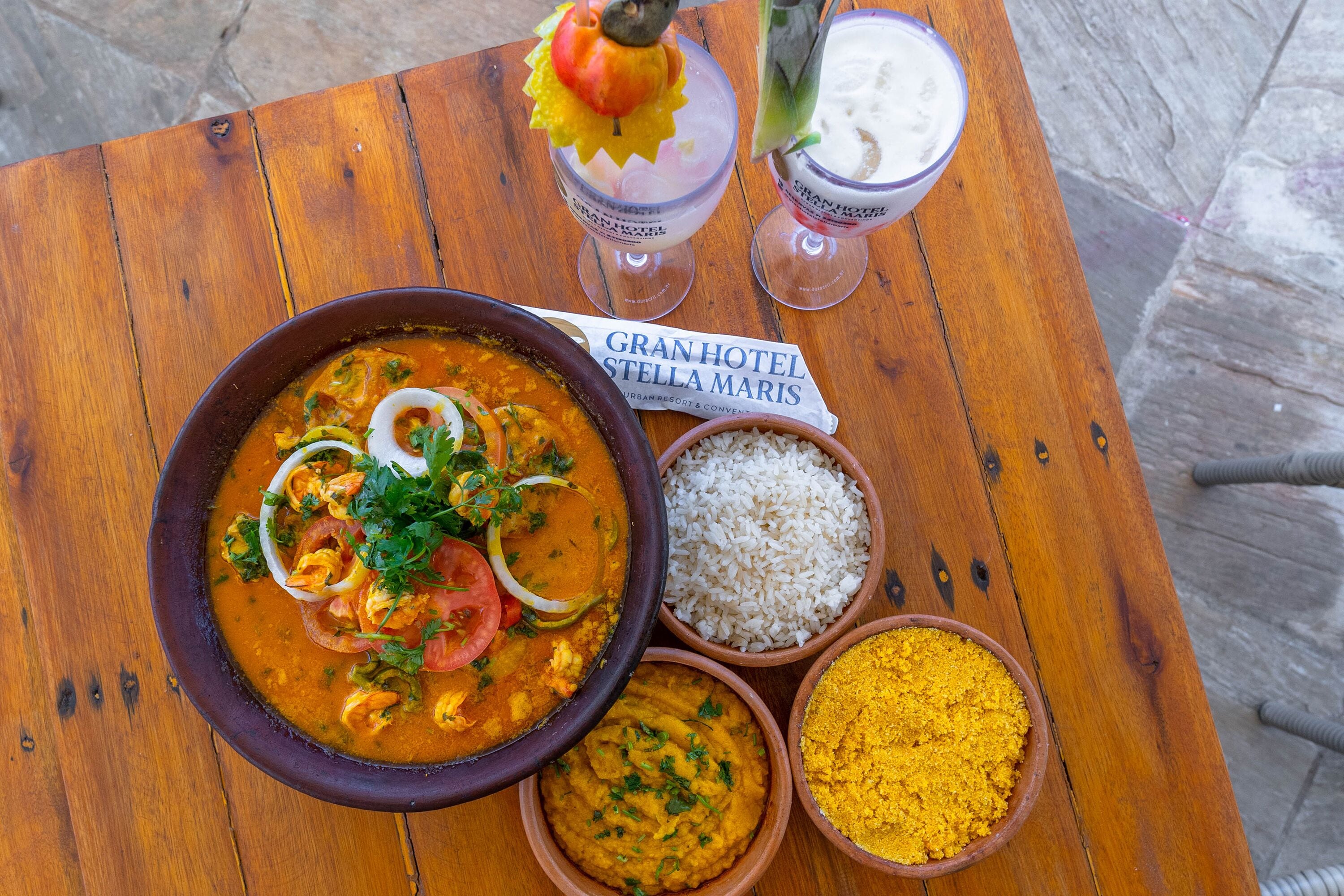 Una mesa de madera con un tazón de curry, arroz, un tazón de especia amarilla y dos bebidas.