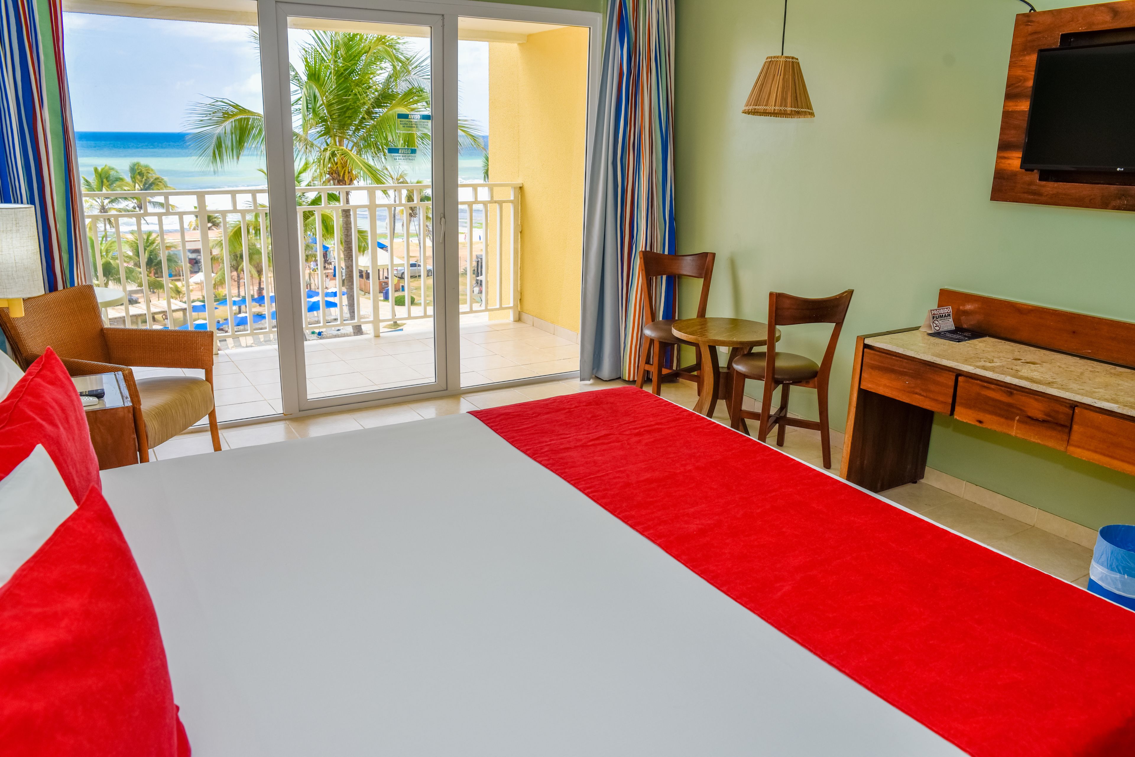 A hotel room with a bed, a desk, a chair, and a view of the beach and palm trees through a sliding door.