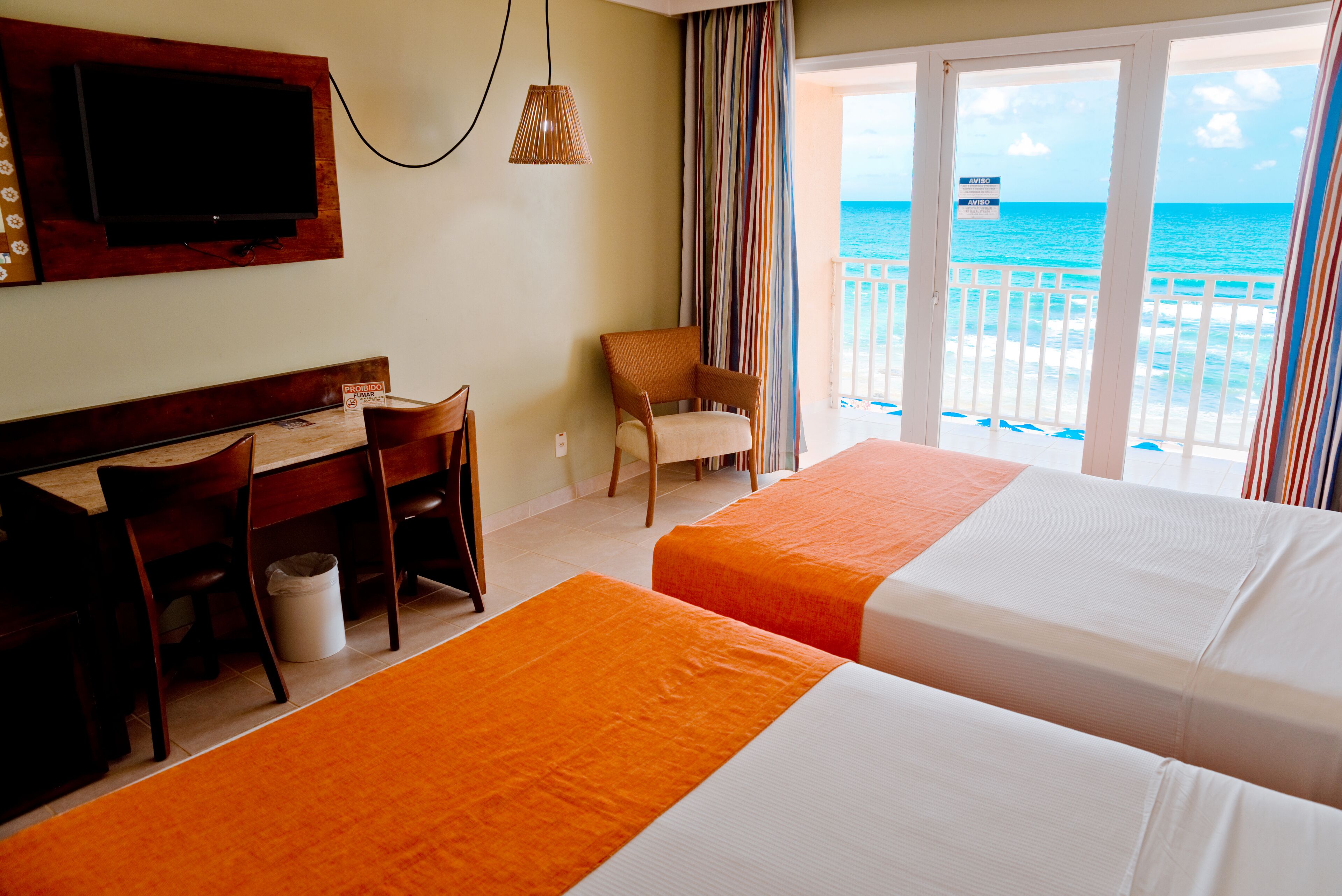 Quarto de hotel com duas camas, uma mesa, uma cadeira e uma TV fixada na parede.