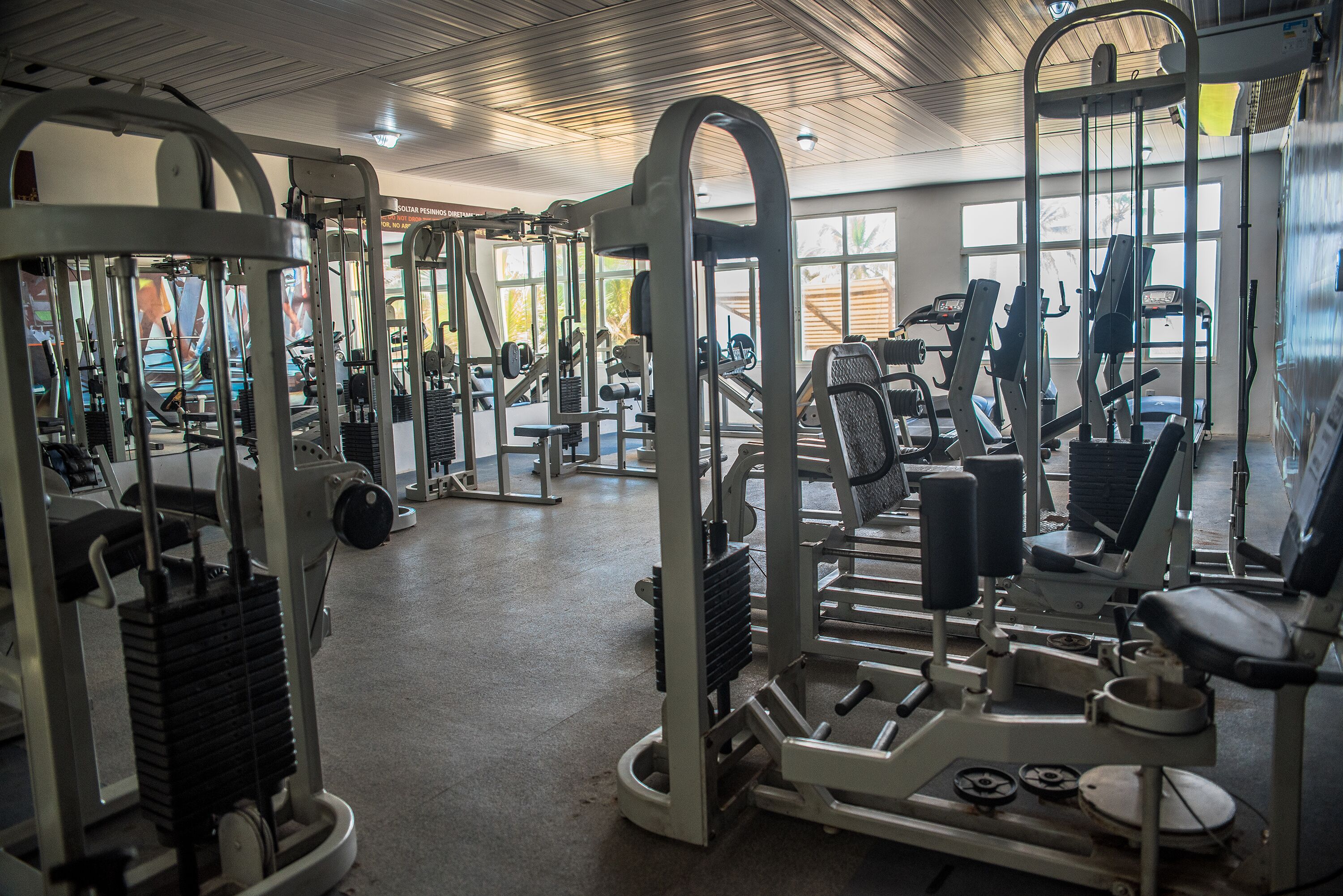 A gym with various exercise machines and equipment, large windows allowing natural light, and a ceiling with a grid pattern.