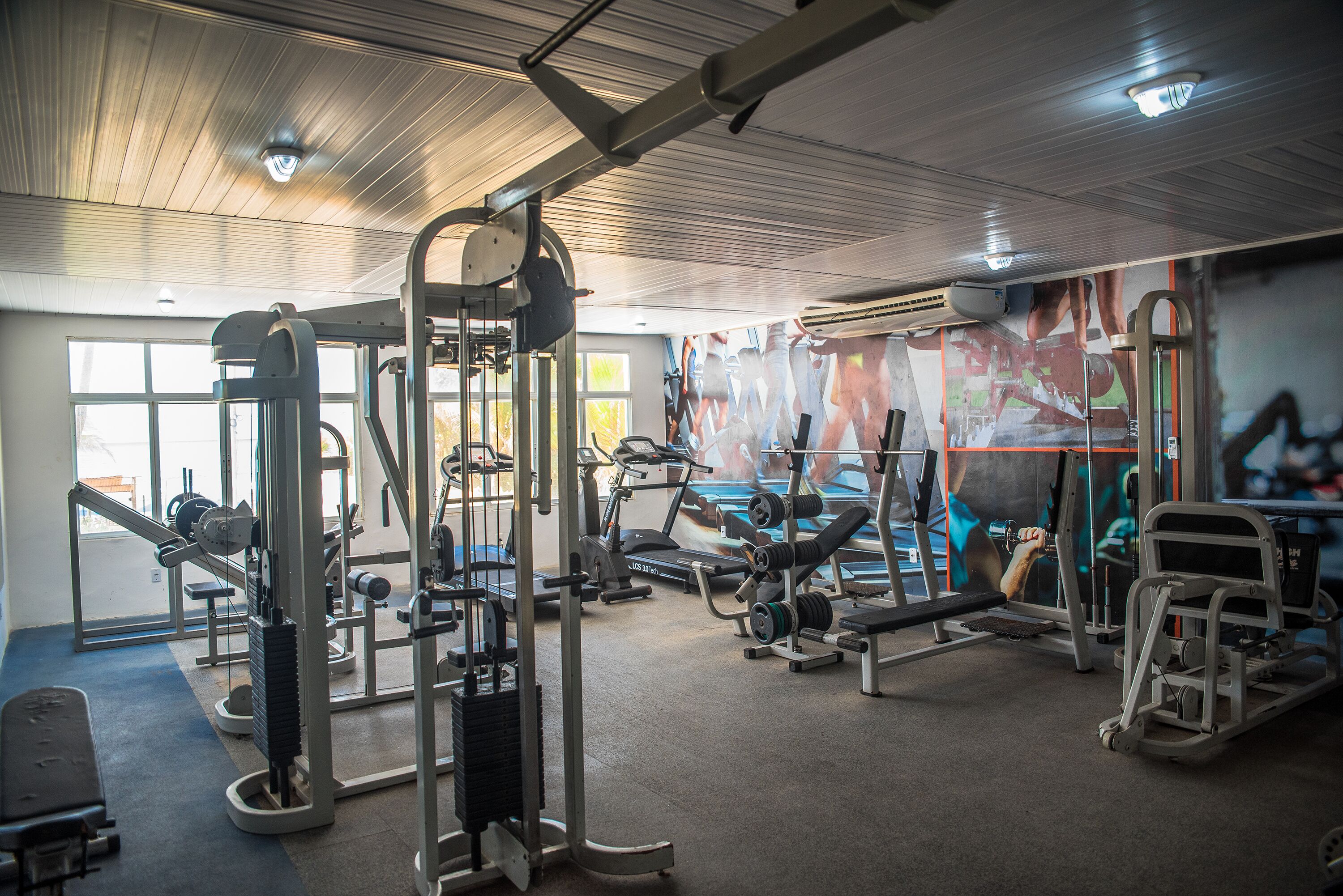A gym with various exercise equipment, large windows, and a mural on the wall.