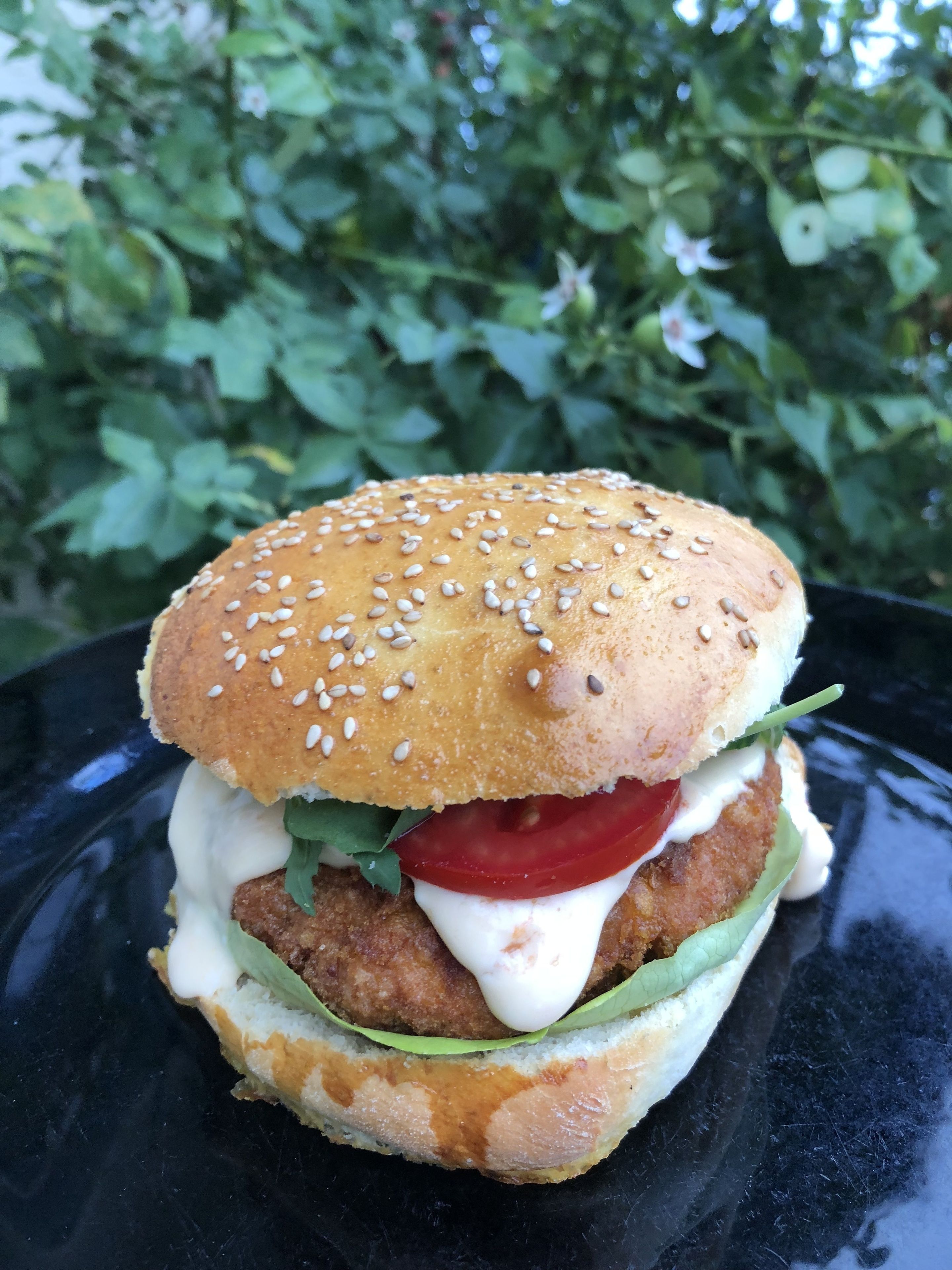 Un sandwich préparé avec un pain aux graines de sésame, de la laitue, des tomates et une galette frite, servi sur une assiette noire.