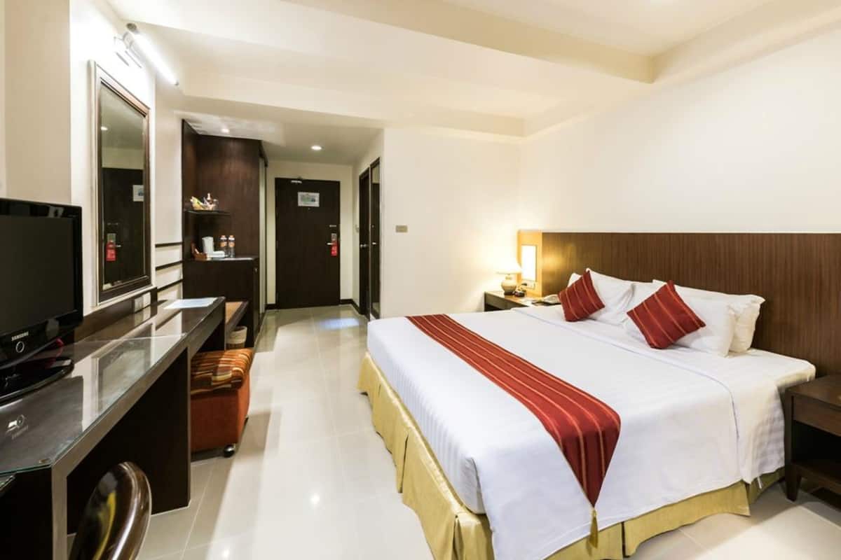 A hotel room with a bed, bedside table, lamp, TV, and a desk with a chair.