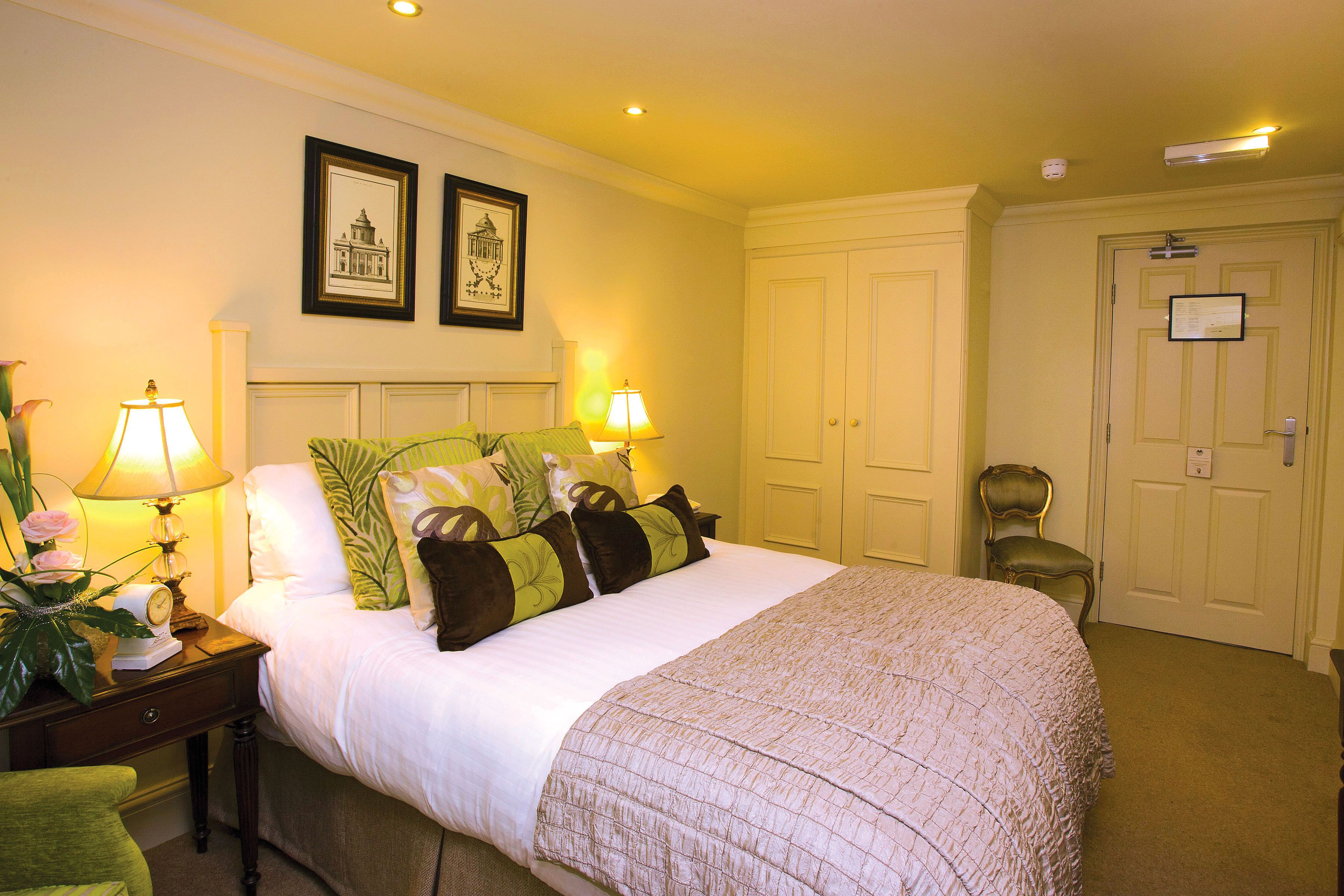 Una cama bien hecha con edredón blanco y almohadones decorativos, una mesita de noche con lámpara y una silla en una habitación de hotel.