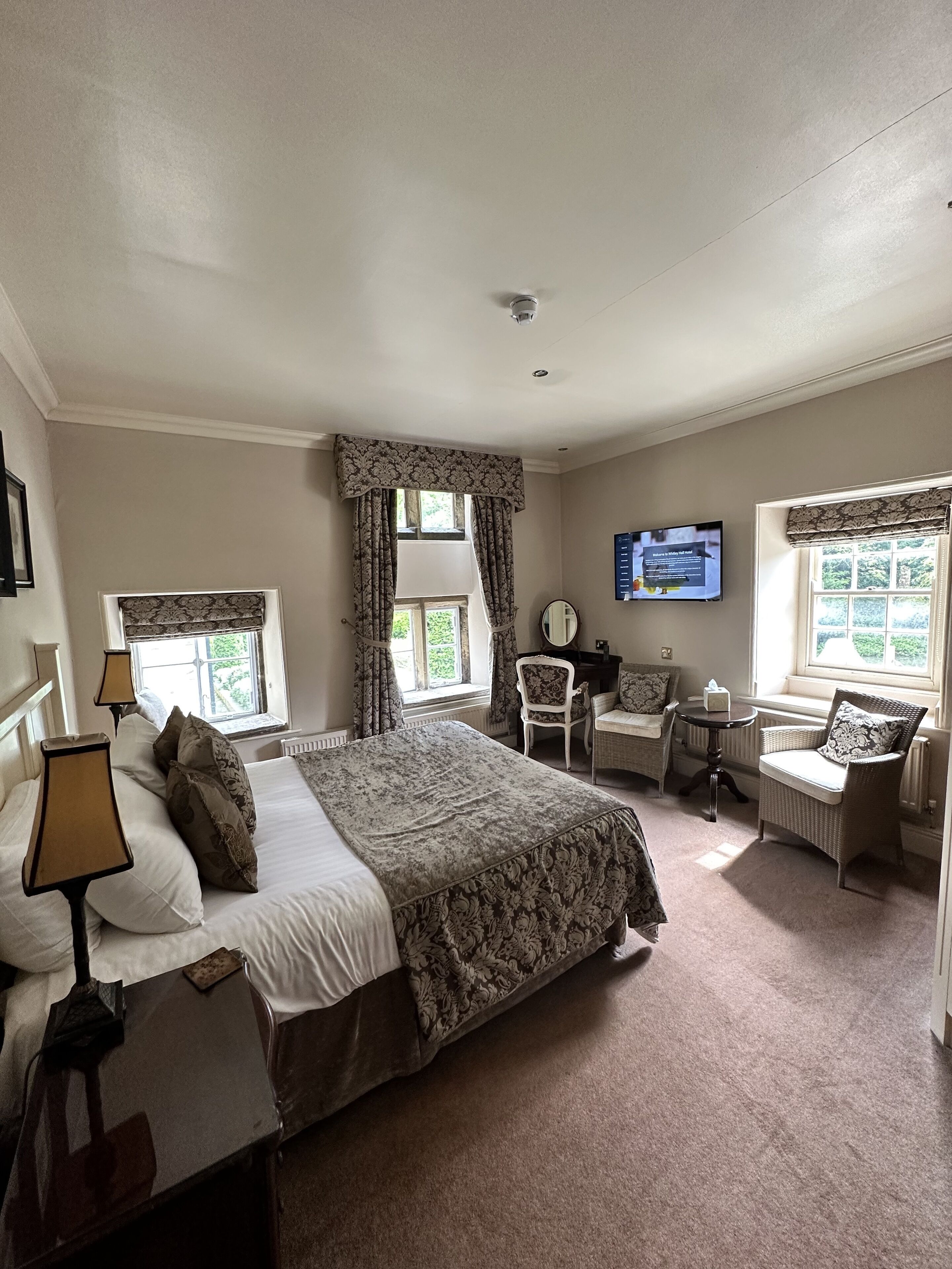 A well-lit bedroom with a large bed, two bedside tables, a desk, a chair, and a television on the wall.