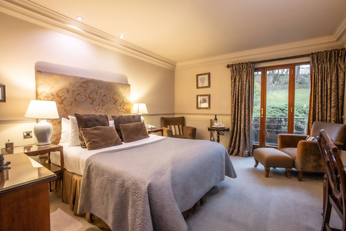Une chambre dotée d’un grand lit, d’un coin salon avec un fauteuil et un pouf, d’une table de chevet avec une lampe, et offrant une vue sur l’extérieur par une fenêtre.