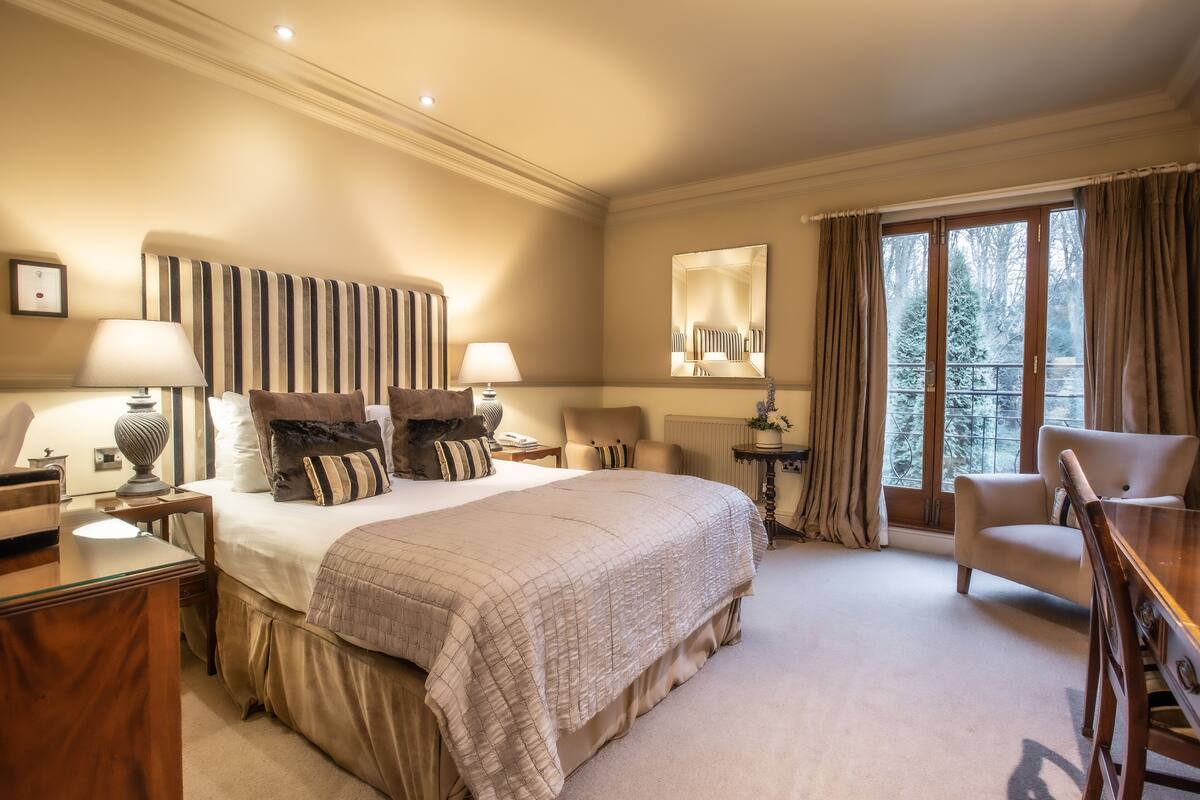 Une chambre d’hôtel avec un grand lit, deux tables de chevet avec des lampes, une chaise, une petite table et une grande fenêtre avec des rideaux.