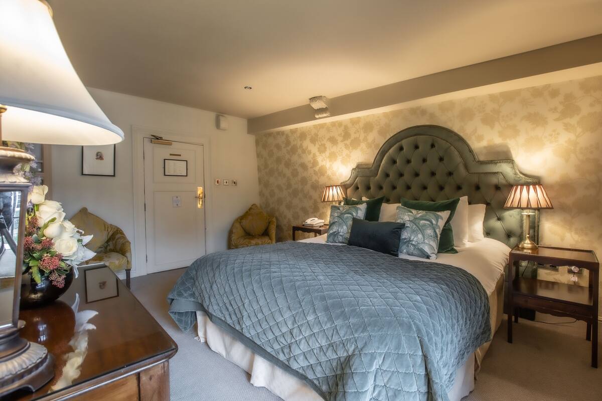 Une chambre à coucher bien rangée, avec un grand lit, deux tables de chevet avec des lampes, une petite table sur laquelle est posé un vase de fleurs et un fauteuil.