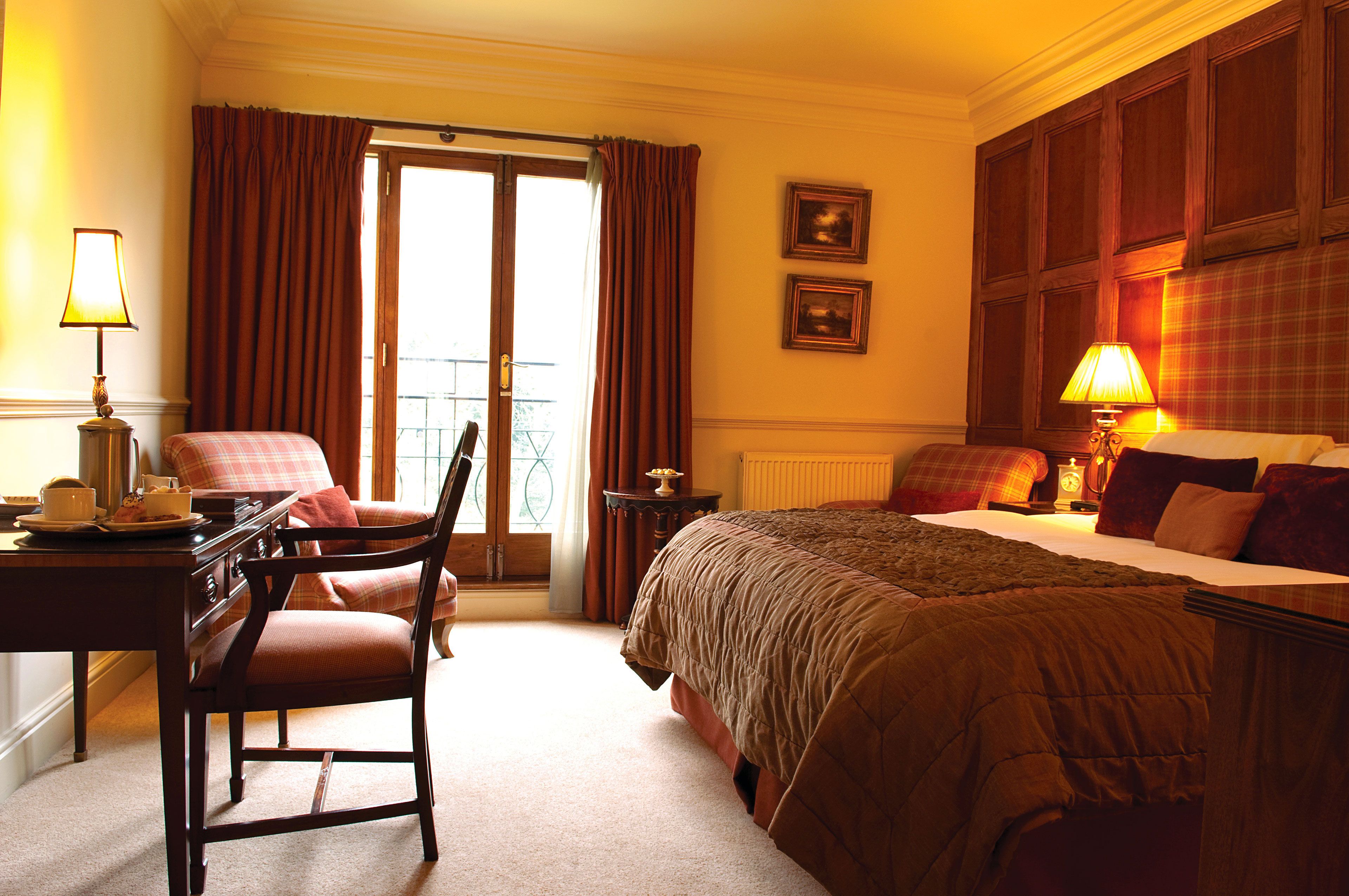 A hotel room with a bed, a chair, a desk with a lamp, a clock, and a window with curtains.
