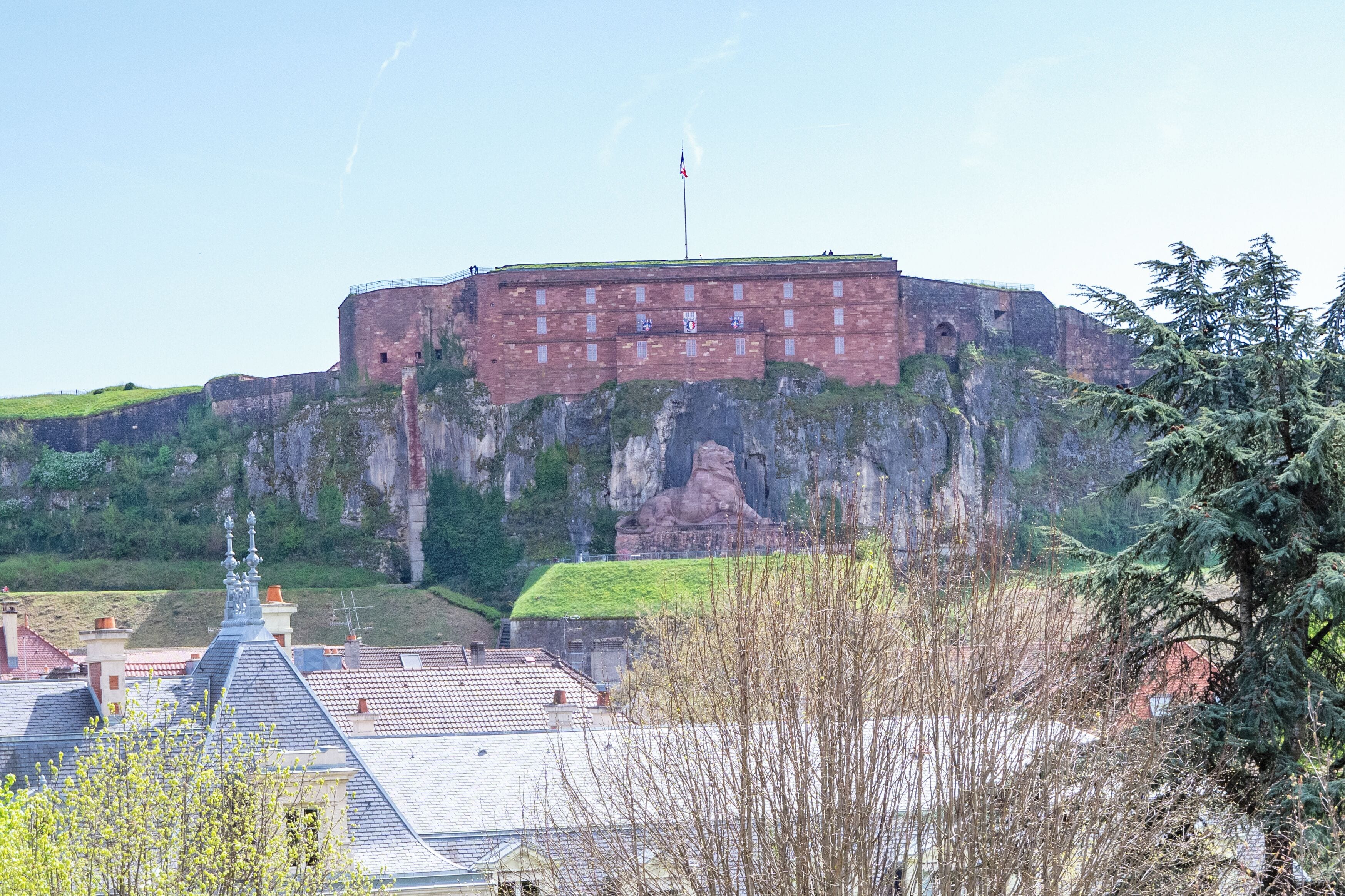 A historic brick building with a flag on top, situated on a cliff with a view of surrounding buildings and greenery.