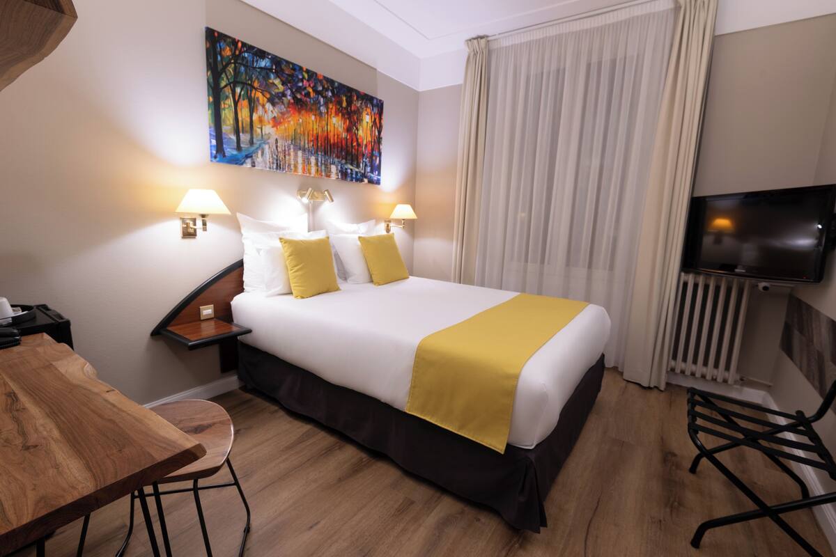 Una habitación de hotel con una cama grande, dos lámparas de noche, un cuadro en la pared, un televisor y un escritorio de madera.