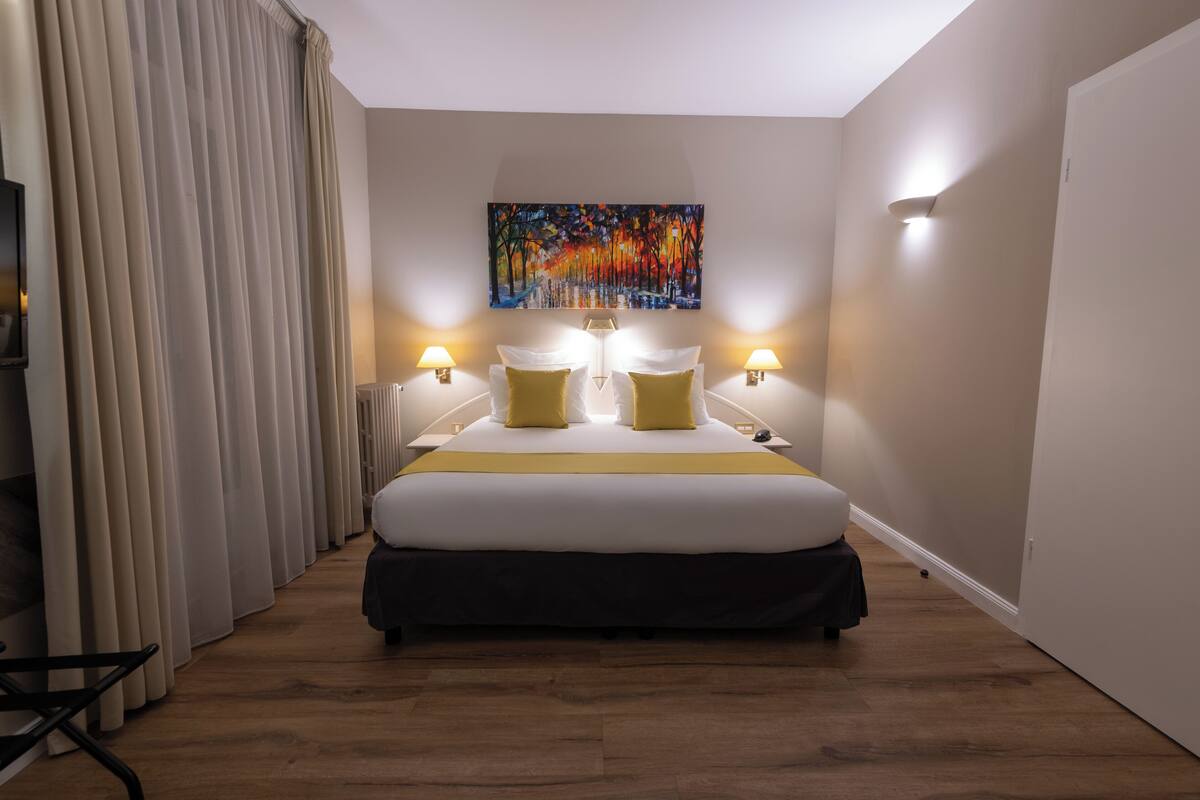 Un cuarto de hotel con una cama grande, dos lámparas de noche, un cuadro en la pared y vistas al exterior a través de la ventana.