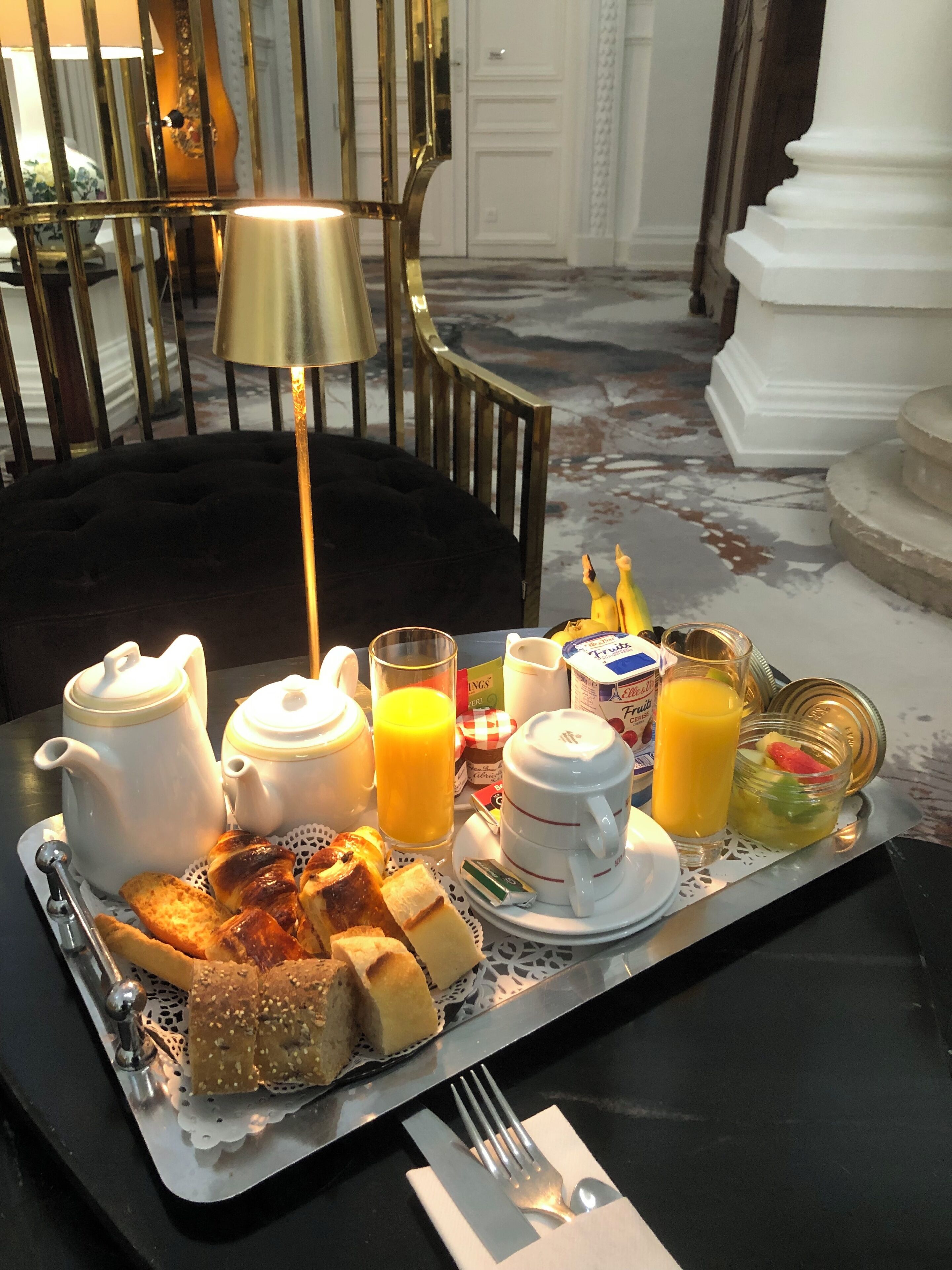 A breakfast tray with croissants, bread, a teapot, a jug of orange juice, a glass of juice, and a fruit bowl.