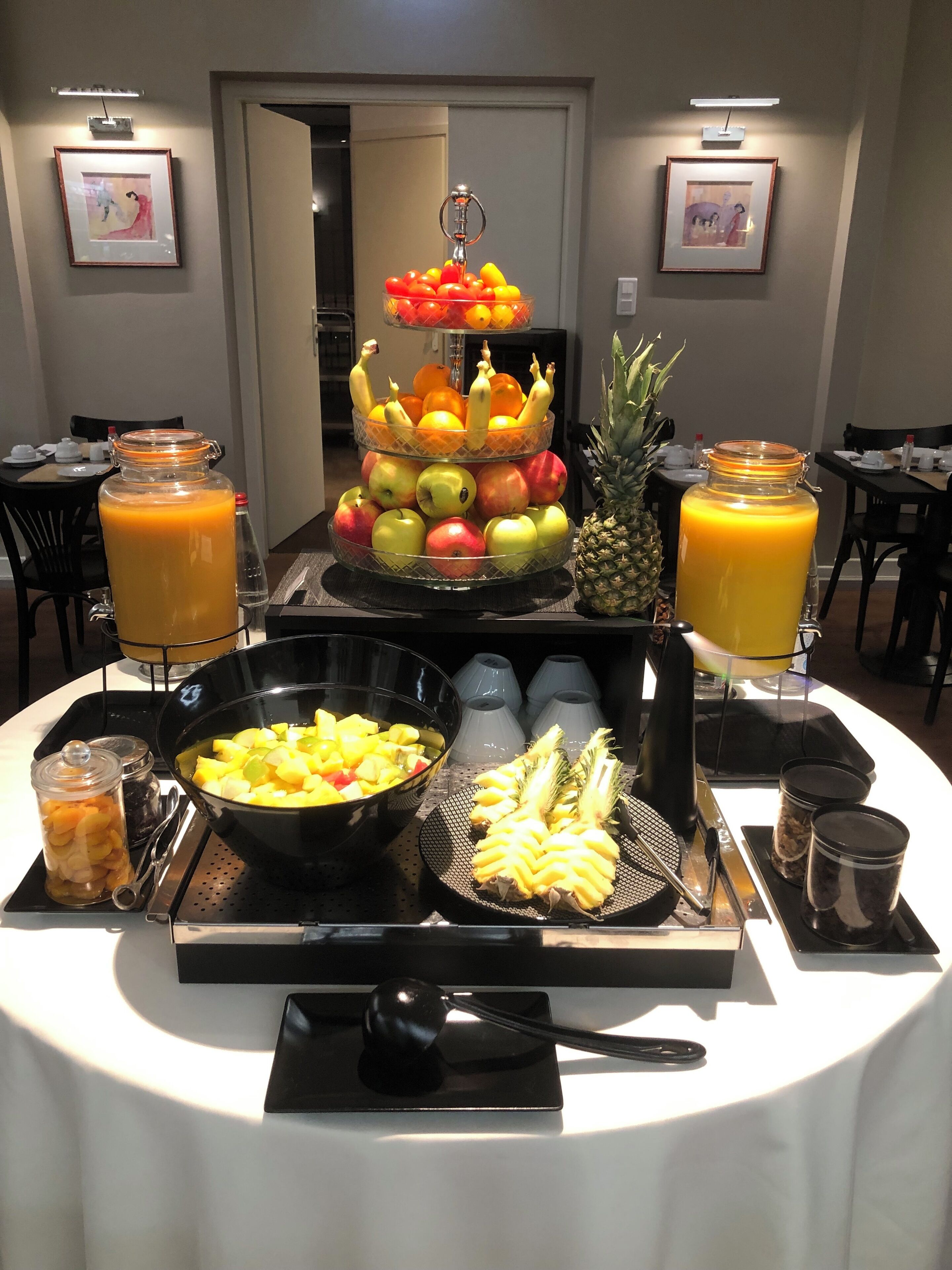 Een buffettafel met een fruitpresentatie in drie verdiepingen, een schaal met ananasstukjes en twee kannen jus d'orange.