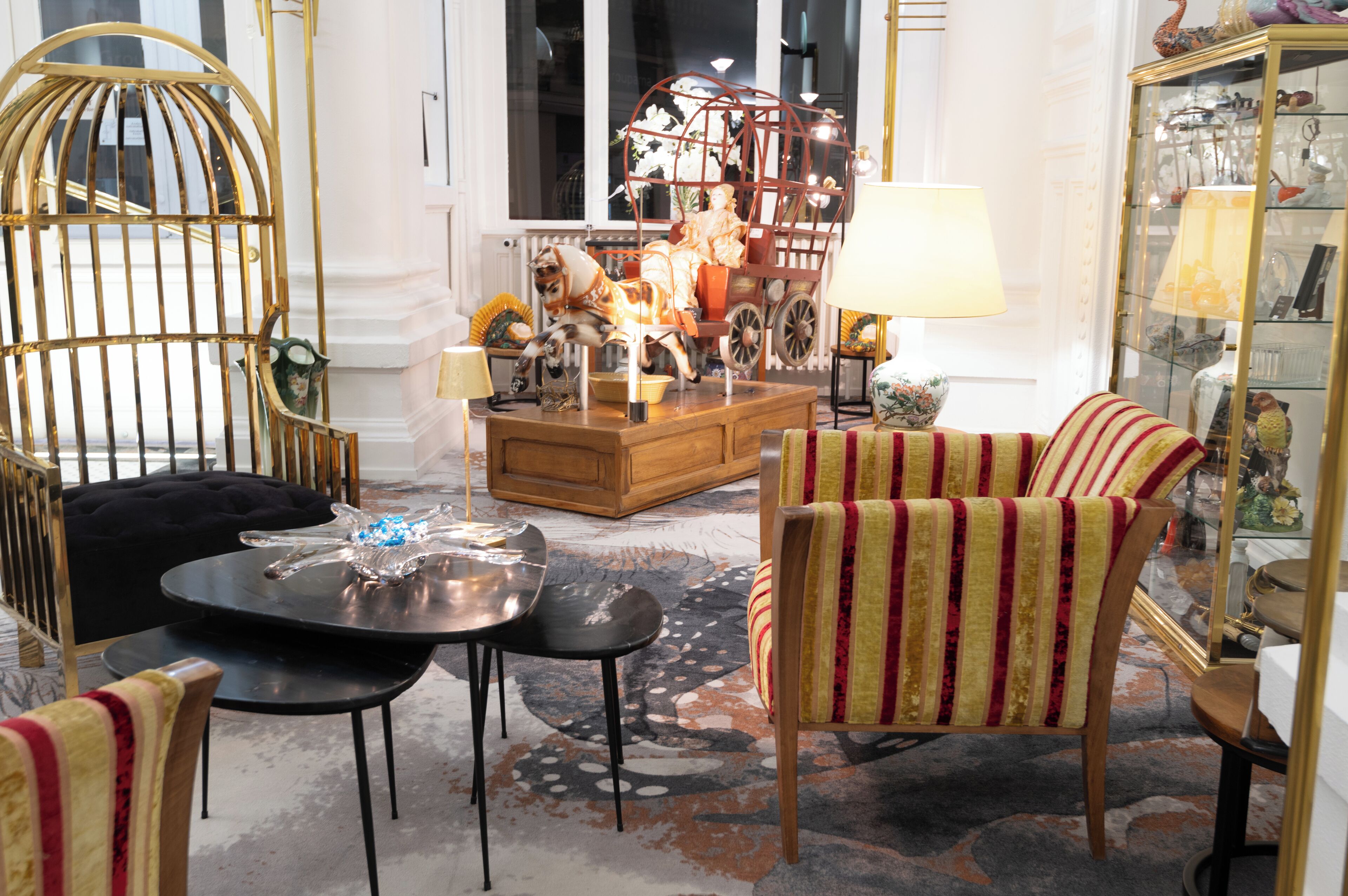 A cozy living room with a wooden coffee table, striped armchairs, a glass cabinet, and a decorative horse carriage.