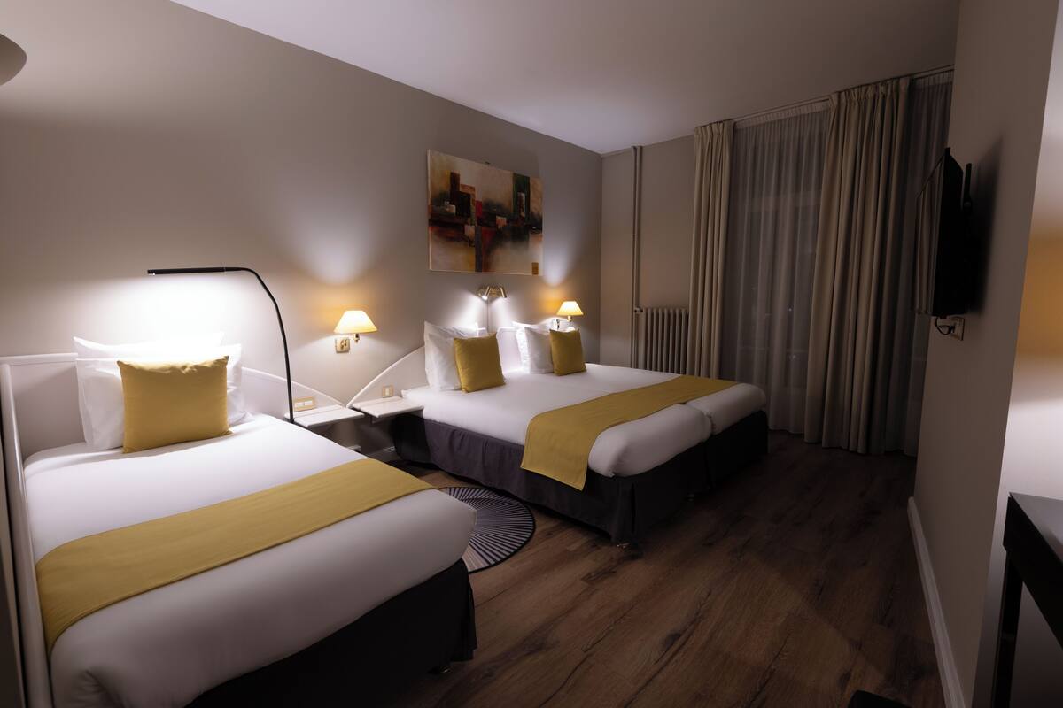 Habitación de hotel con dos camas, cada una con ropa de cama blanca y detalles amarillos, una mesita de noche con lámparas, un cuadro grande en la pared y vistas de la ciudad a través de cortinas traslúcidas.