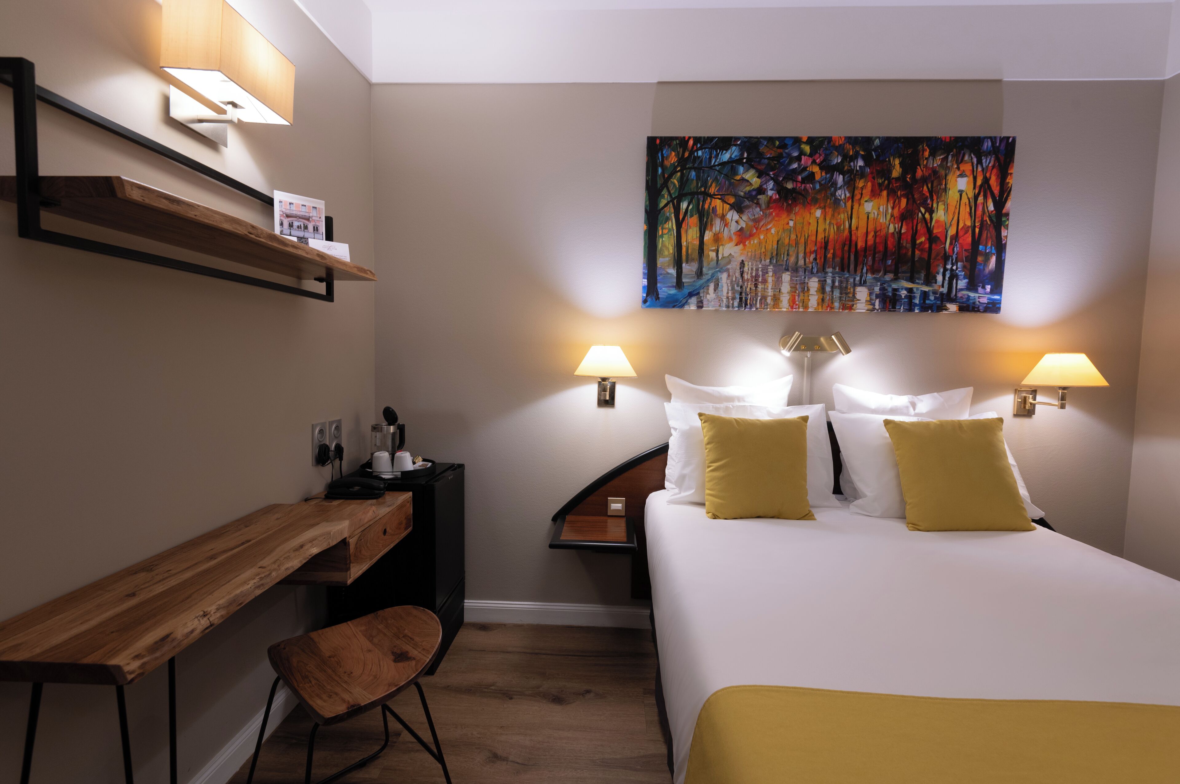 A hotel room with a bed, bedside lamps, a desk, a chair, a painting on the wall, and a shelf with a lamp.