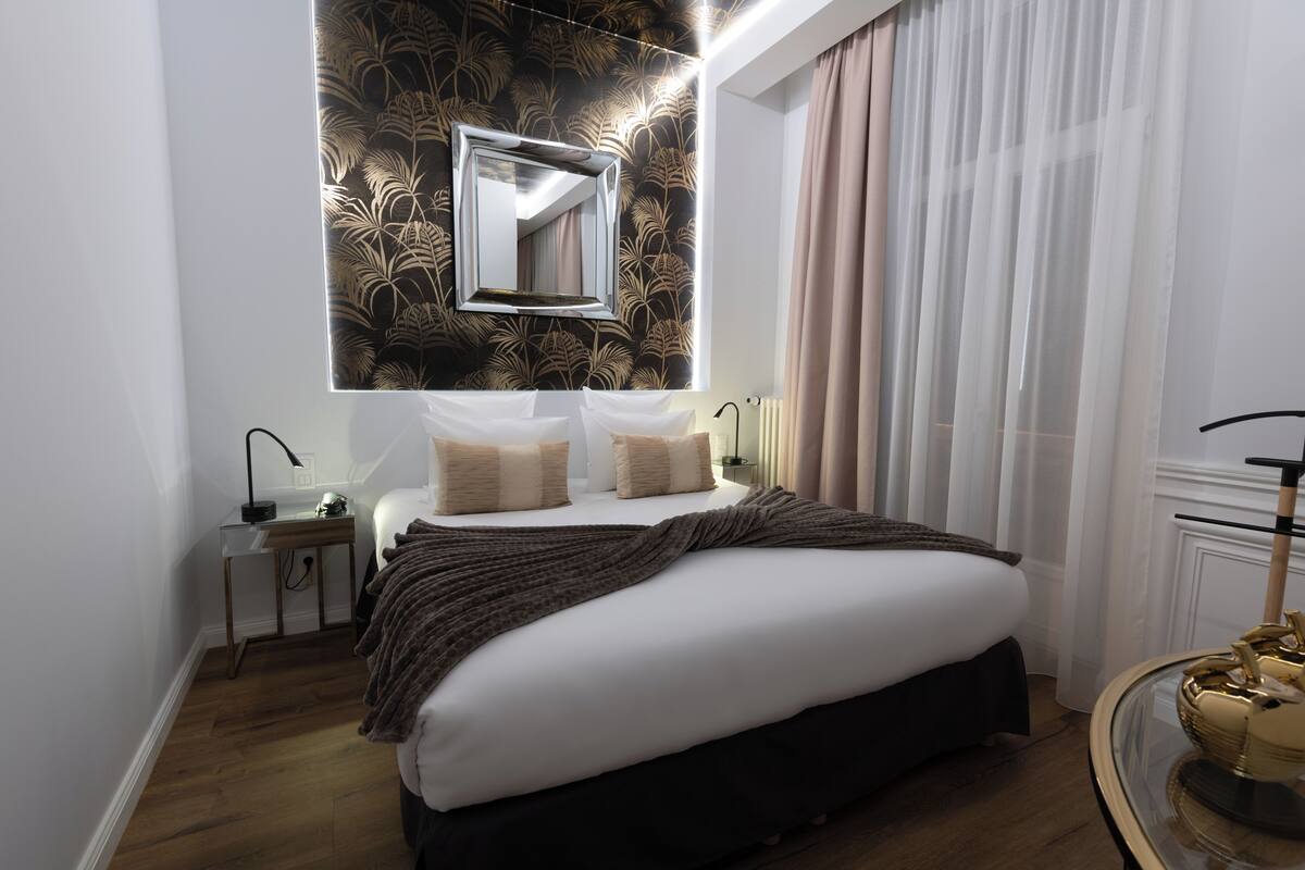 Una habitación de hotel moderna con una cama grande, una mesita de noche, un espejo y una ventana con cortinas.