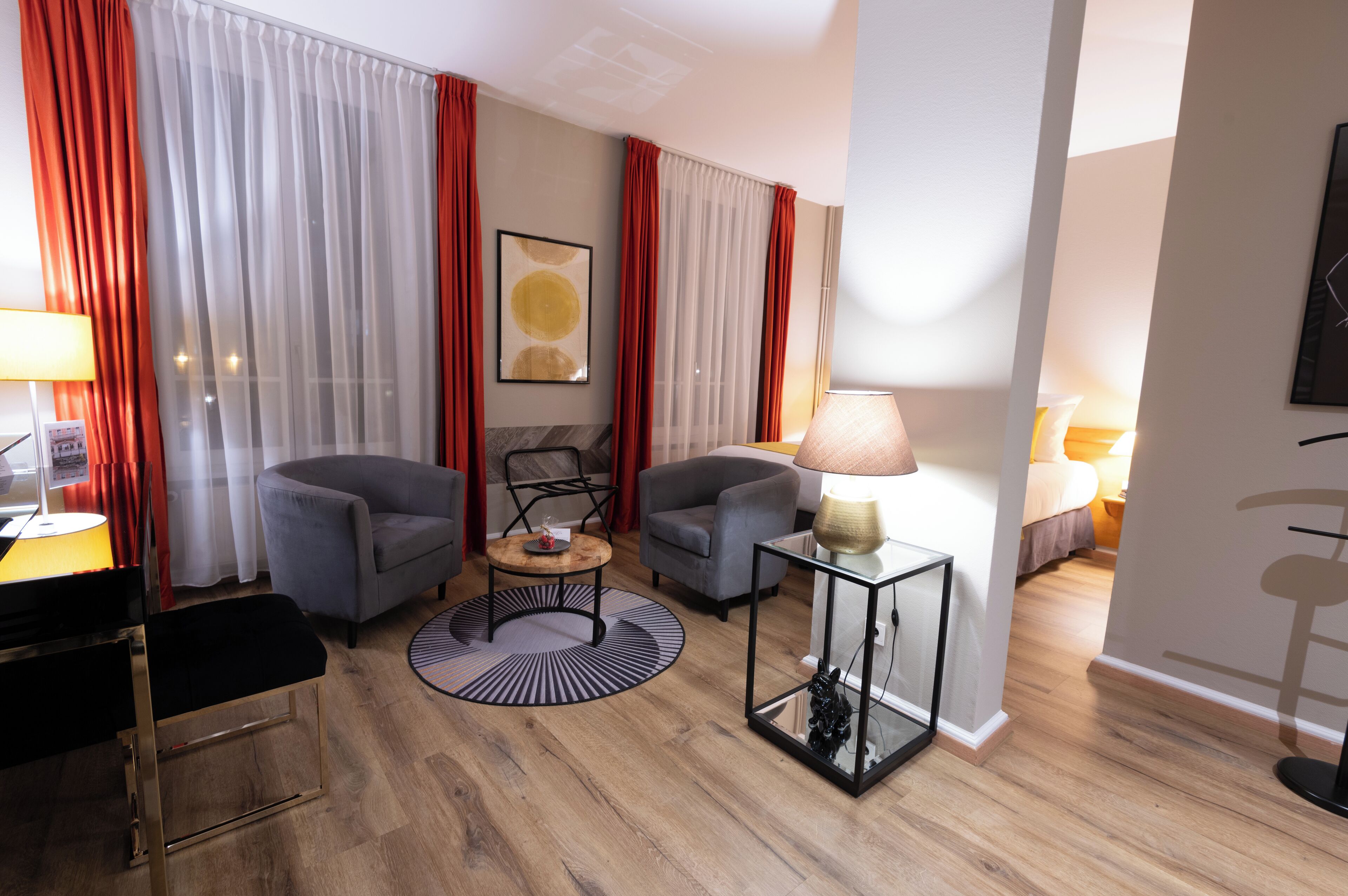 Una habitación de hotel moderna con dos sillones grises, una mesita redonda pequeña y una mesa de centro de vidrio, además de una cama con cabecera blanca.