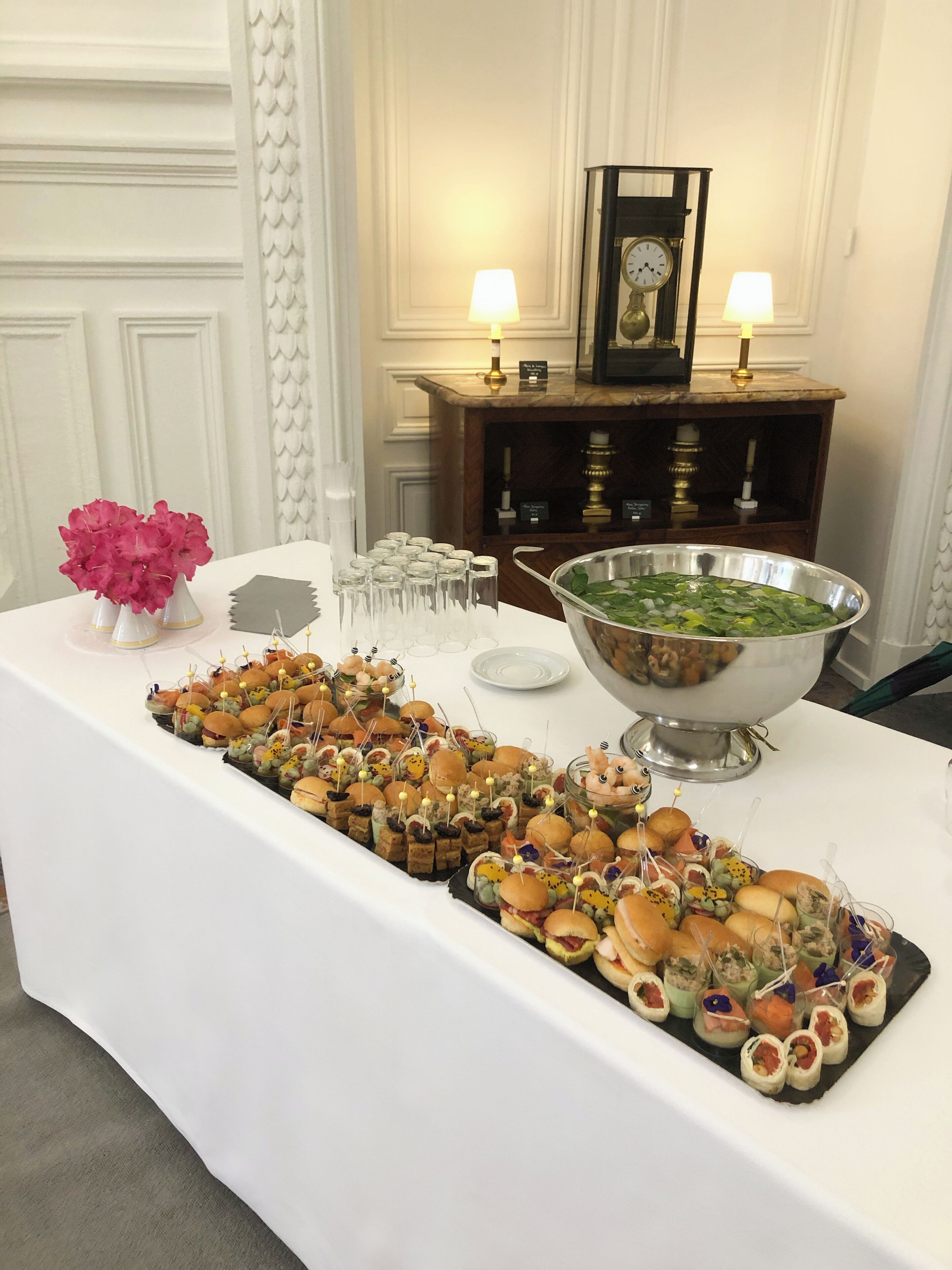 A buffet table with various food items, a large bowl of salad, and a clock on a wooden cabinet in the background.