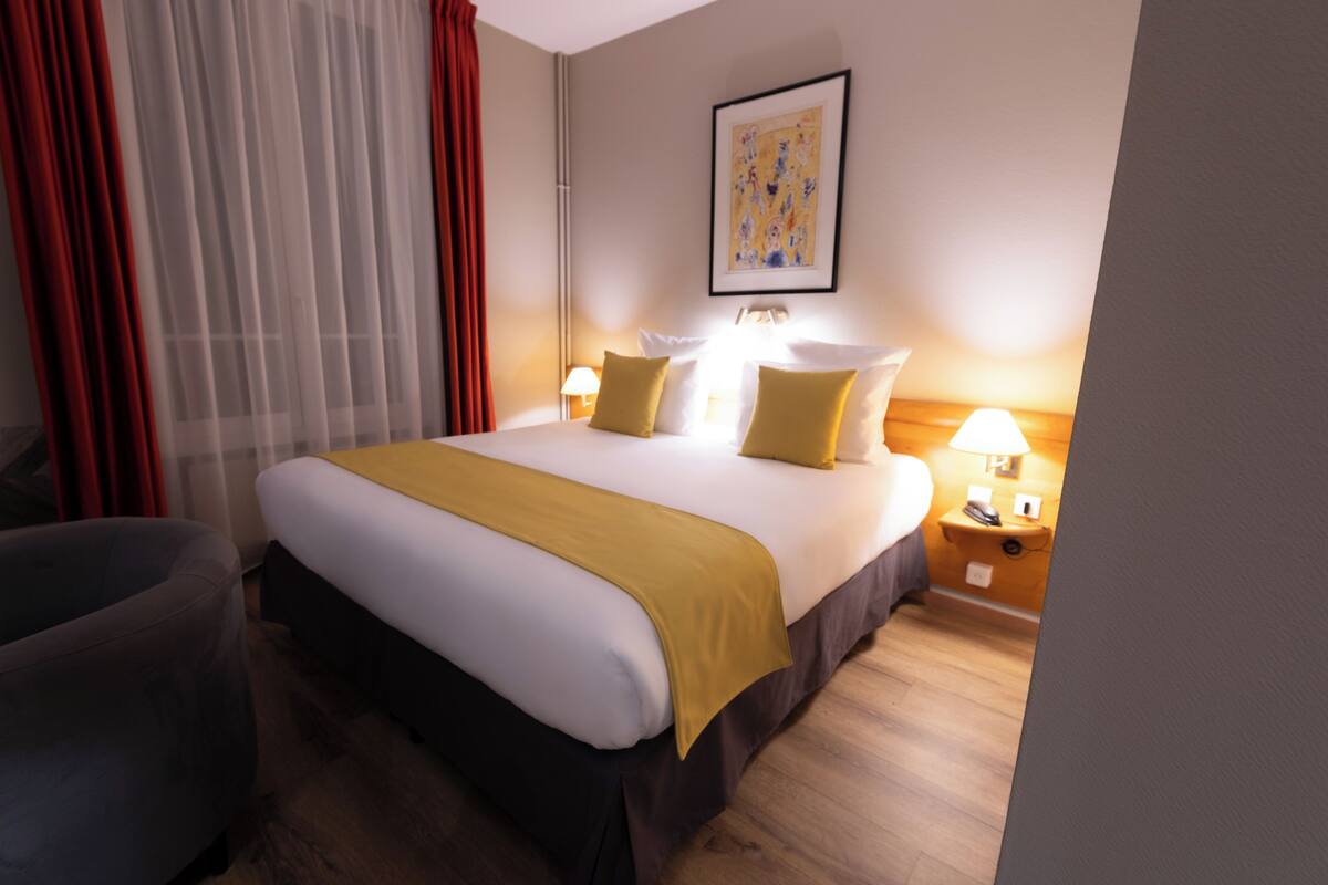 Habitación de hotel con cama, dos mesitas de noche, una silla y una mesita con lámpara.