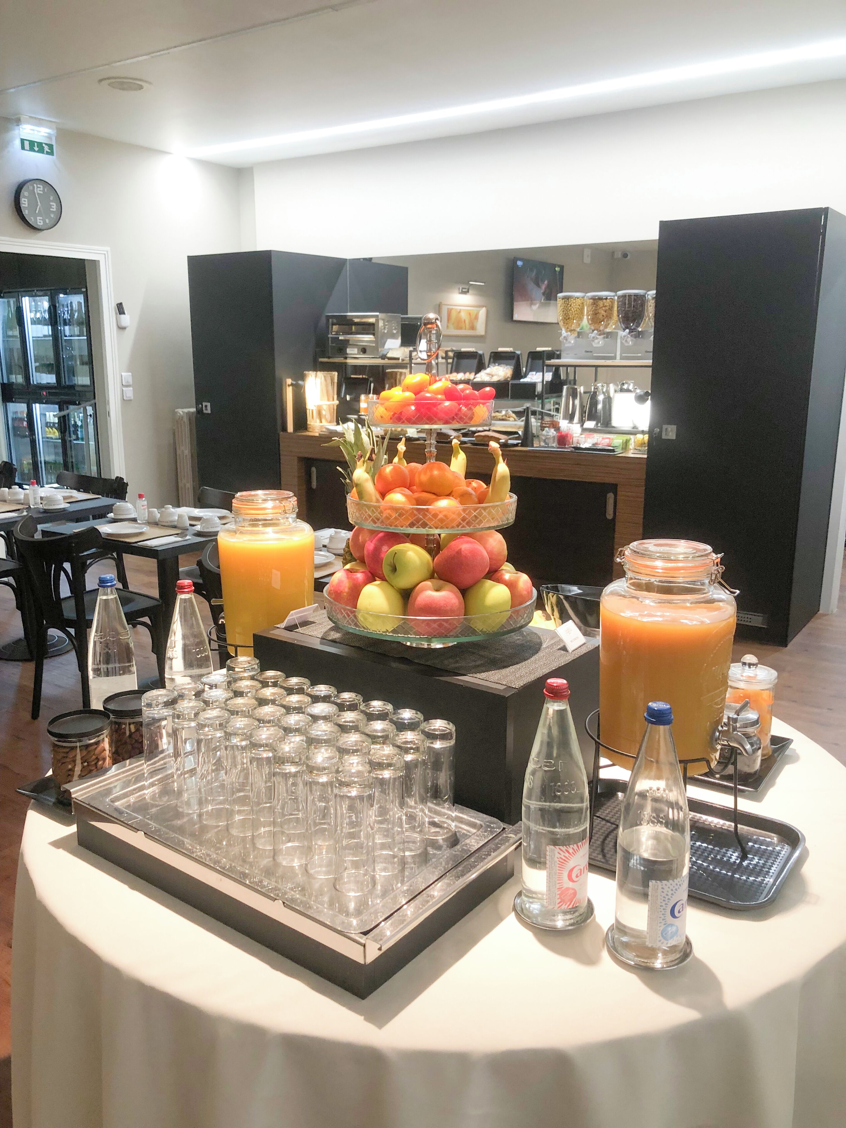 A buffet setup with a fruit display, beverage dispensers, and a clock on the wall.