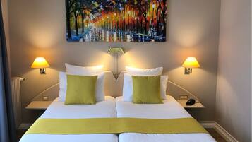 Uma cama de casal com roupa de cama branca e verde, duas luminárias de cabeceira, um quadro acima da cama e piso de madeira.