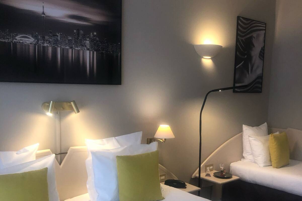 Una habitación de hotel con dos camas, cada una con ropa blanca y almohadas verdes, una mesita de noche con una lámpara y un cuadro de un paisaje urbano en la pared.