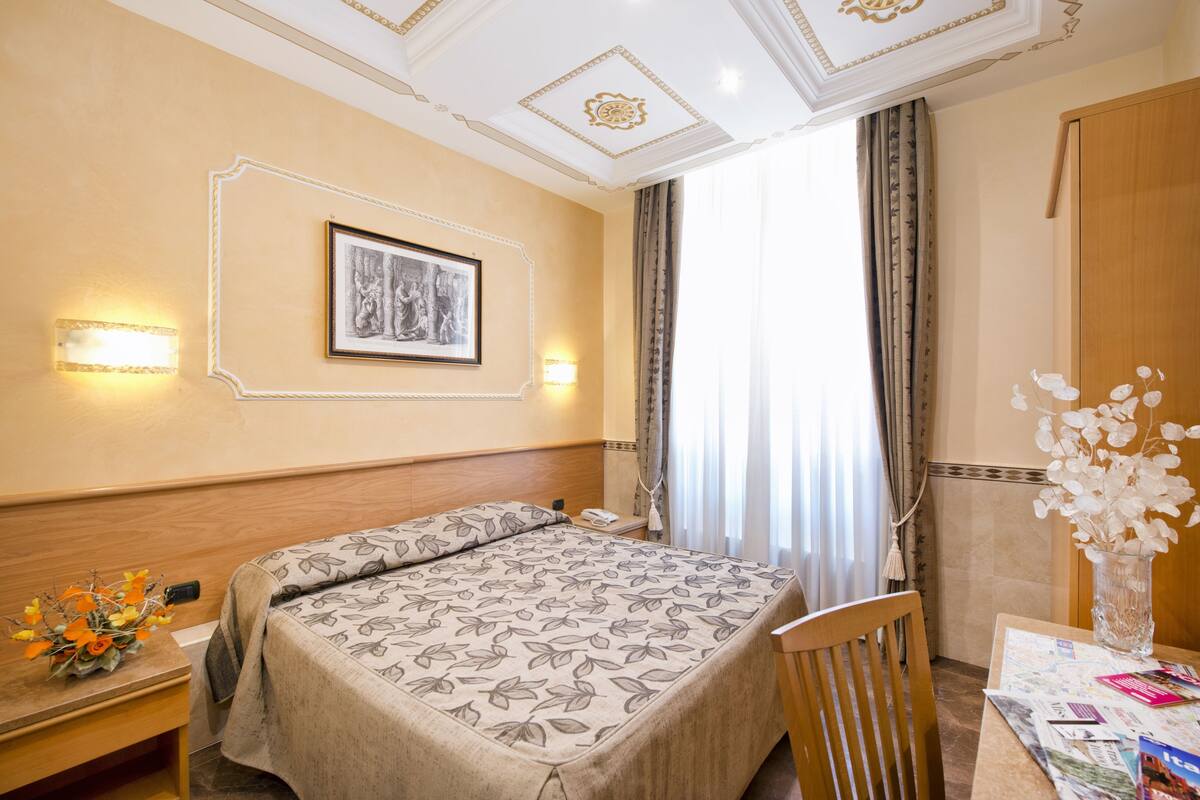 A hotel room with a bed, bedside table, lamp, framed artwork, and a vase with flowers.
