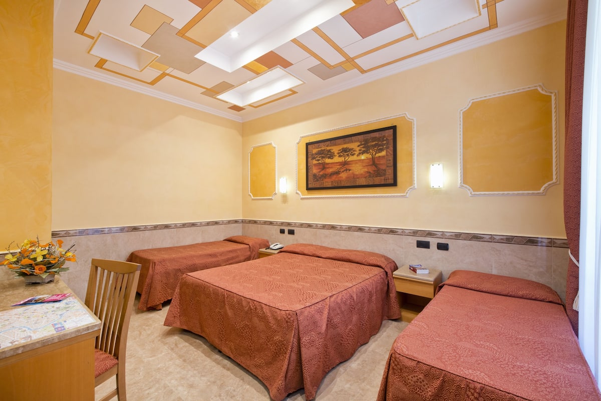 A hotel room with two beds, a desk, and a chair. There is a painting on the wall and a vase with flowers on the desk.