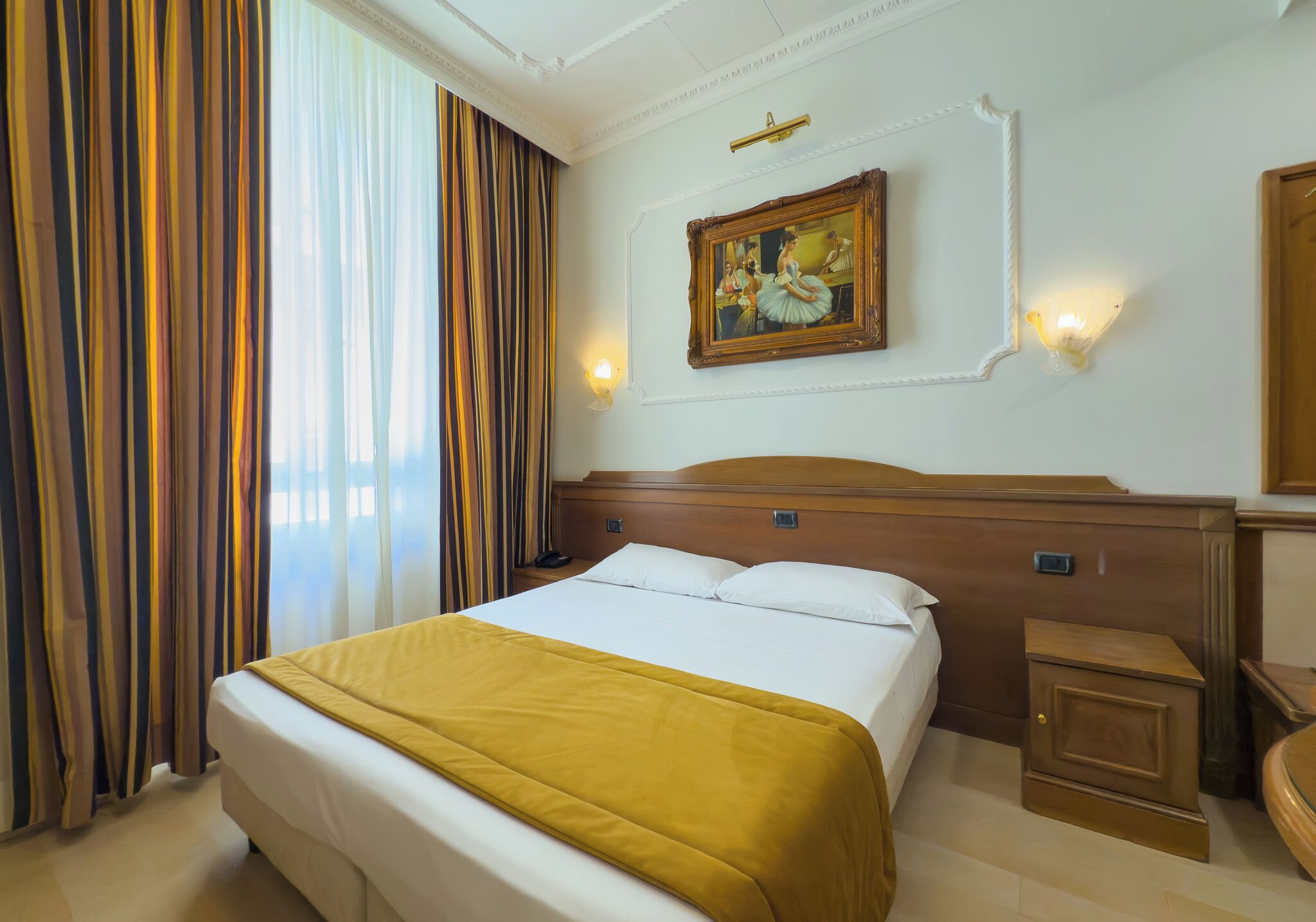 A hotel room with a large bed, a nightstand, a wooden headboard, a painting on the wall, and a window with curtains.