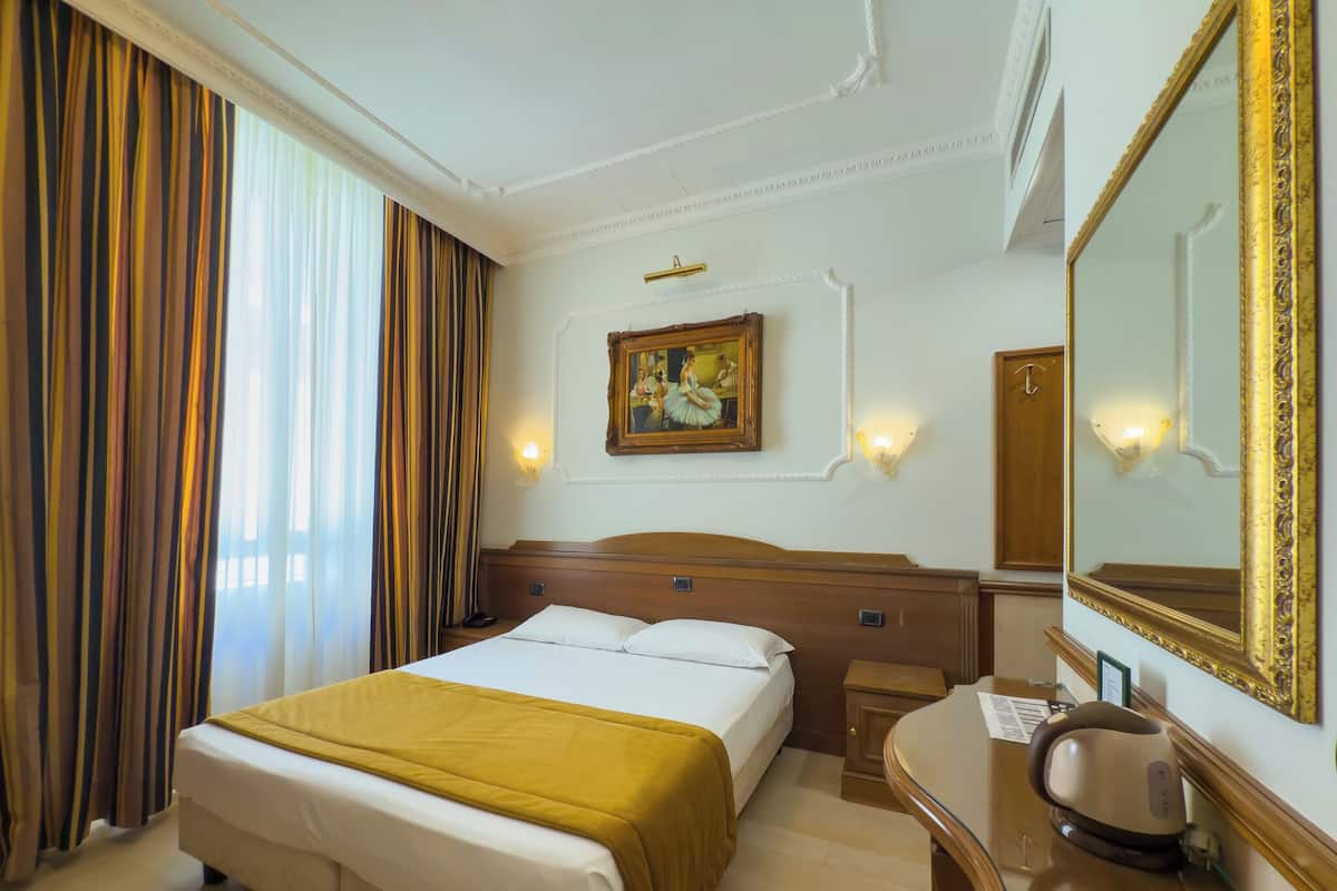 A hotel room with a bed, a desk, a chair, a large mirror, a painting on the wall, and a window with curtains.