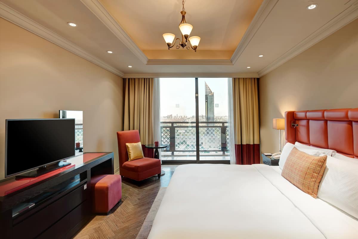 A hotel room with a large bed, a TV on a desk, a red chair, and a balcony with a city view.