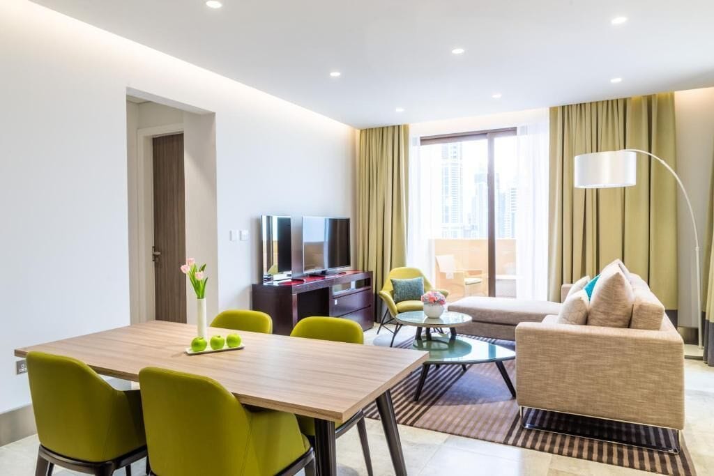 A modern hotel room with a dining area, a sofa, a TV, and a large window with curtains.