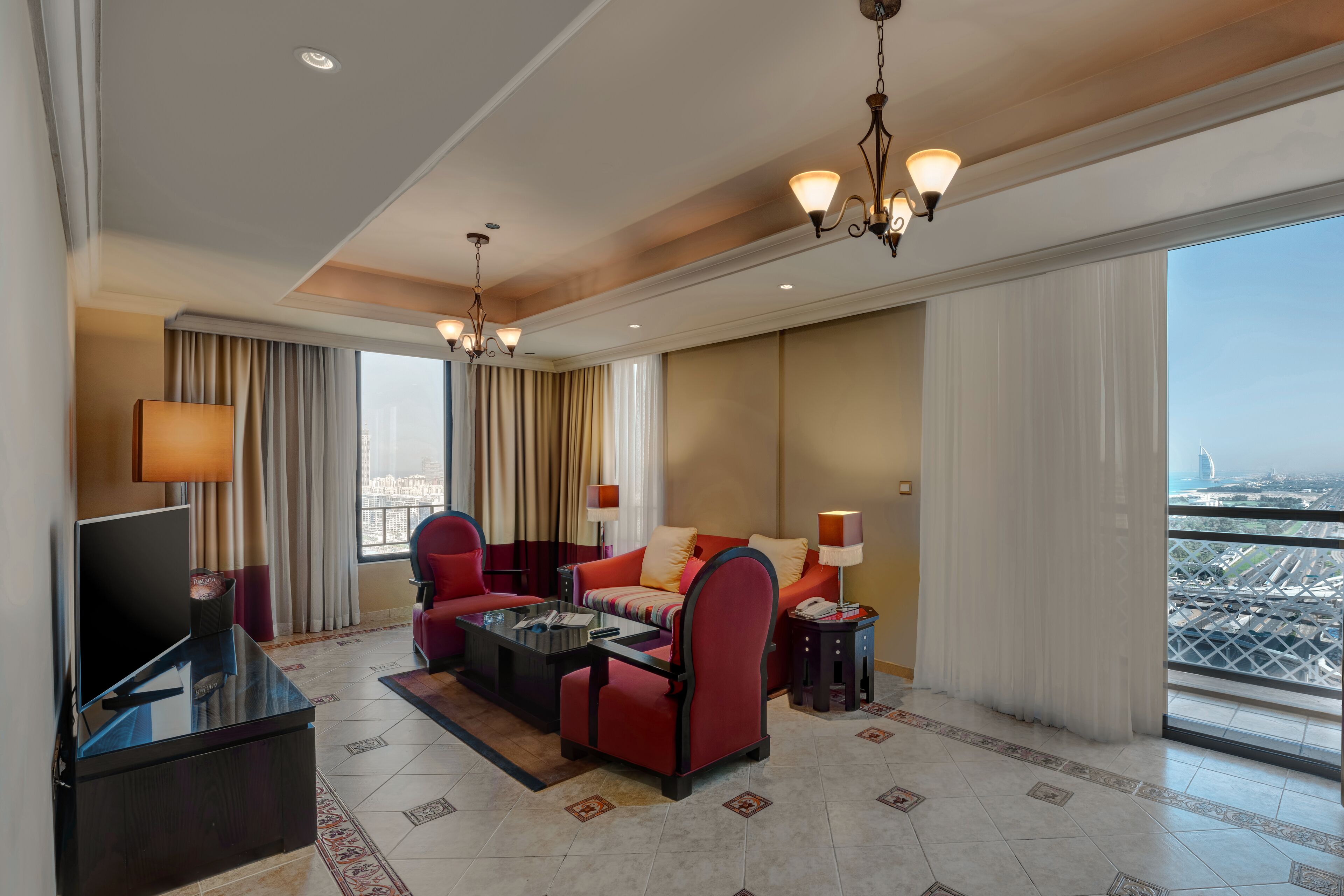 A spacious hotel room with a large bed, a sitting area with a red sofa and armchairs, a flat-screen TV, and a city view through large windows.