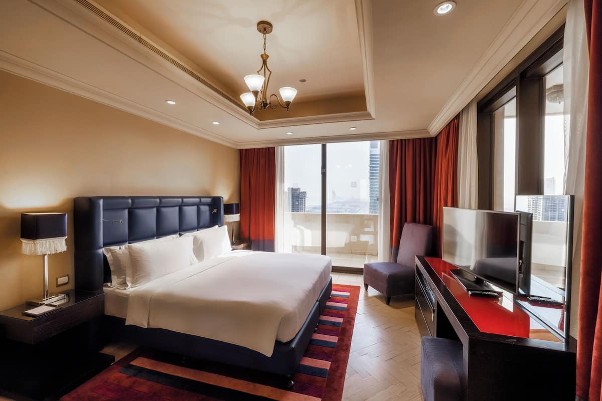 A hotel room with a large bed, a TV, a desk, and a balcony with a city view.