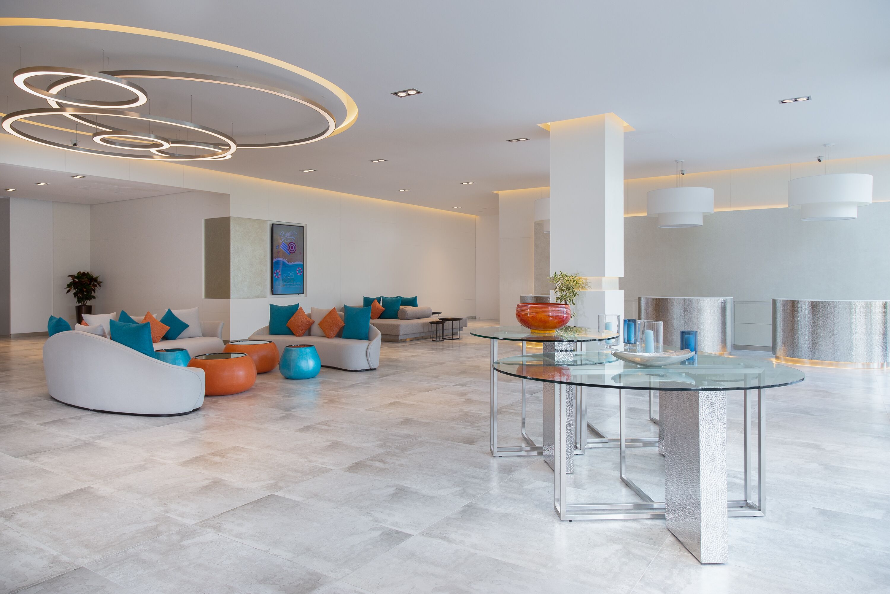 Eine moderne Lobby mit einem Glastisch, weißen Sofas mit bunten Kissen und einer zeitgemäßen Deckenleuchte.