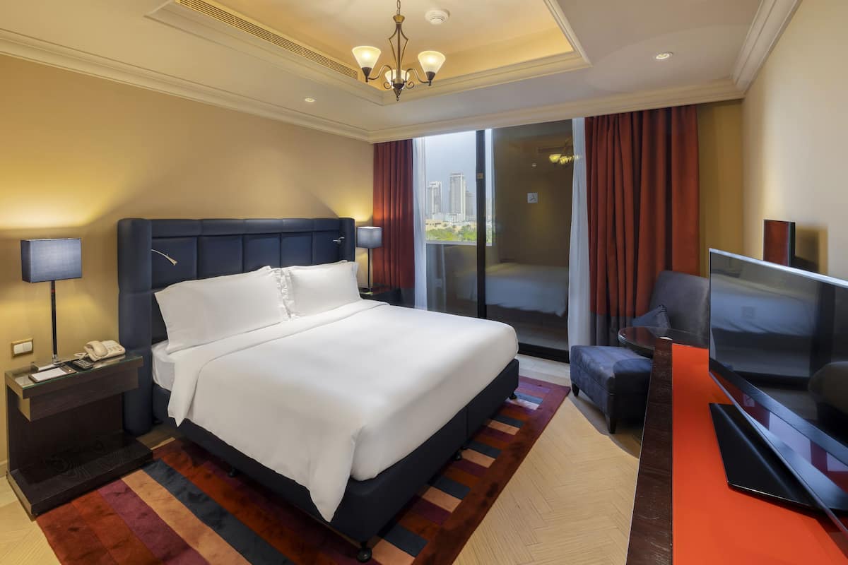 A modern hotel room with a large bed, a TV, a desk with a lamp, and a view of the city through the window.