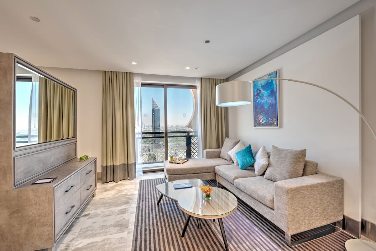 A modern hotel room with a sofa, a glass coffee table, a dresser, and a large window offering a city view.