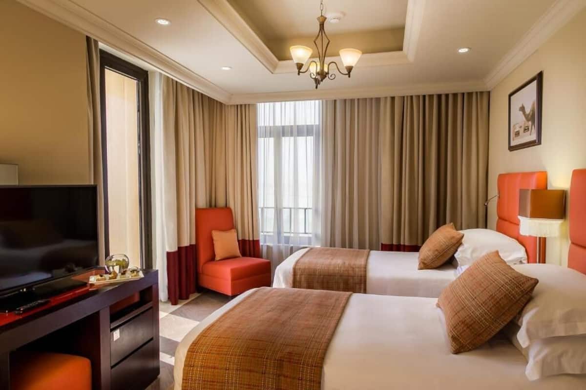 A hotel room with two beds, a TV, a desk, and a large window with curtains.