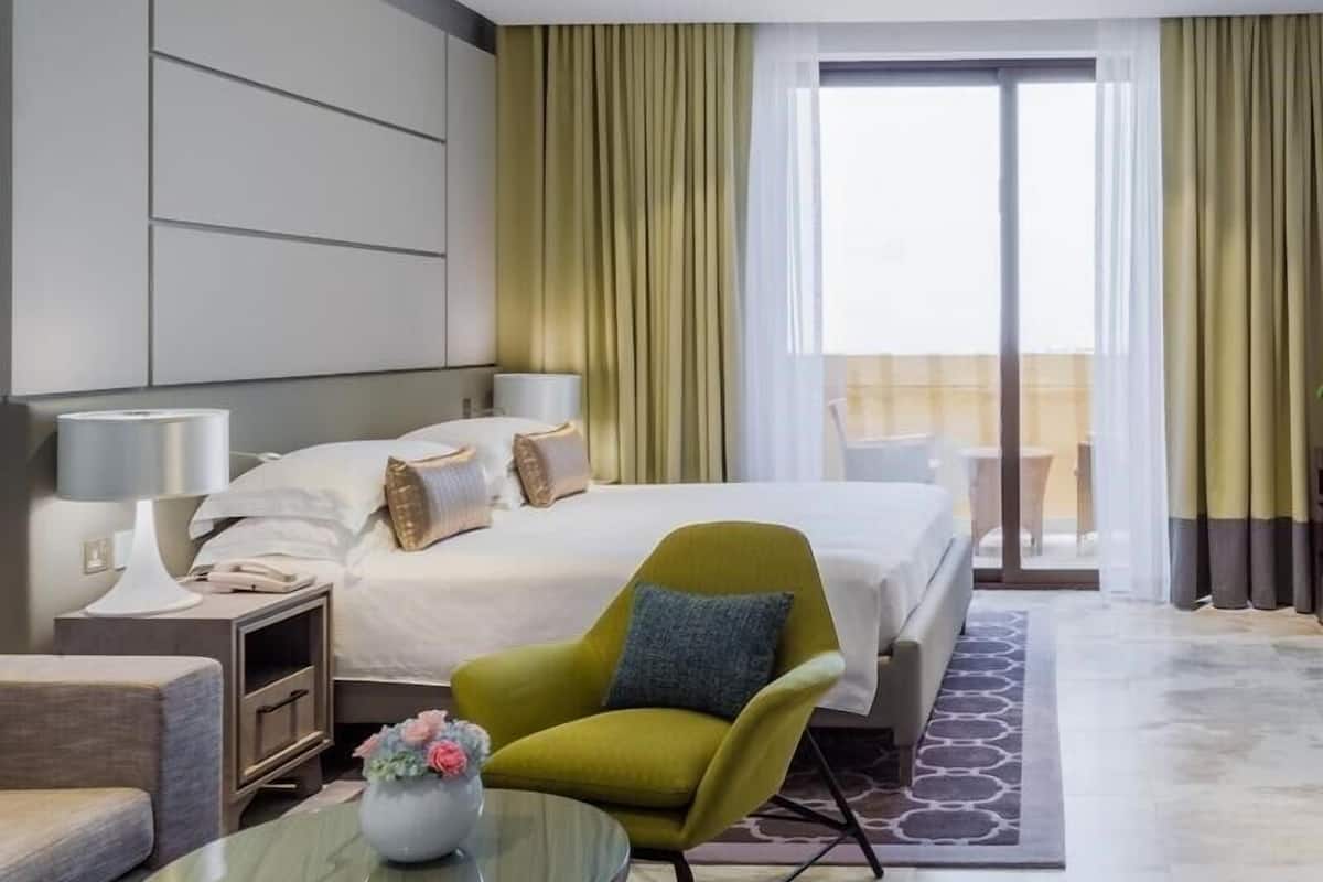 A modern hotel room with a large bed, a green armchair, a glass coffee table, and a view of the outdoors.