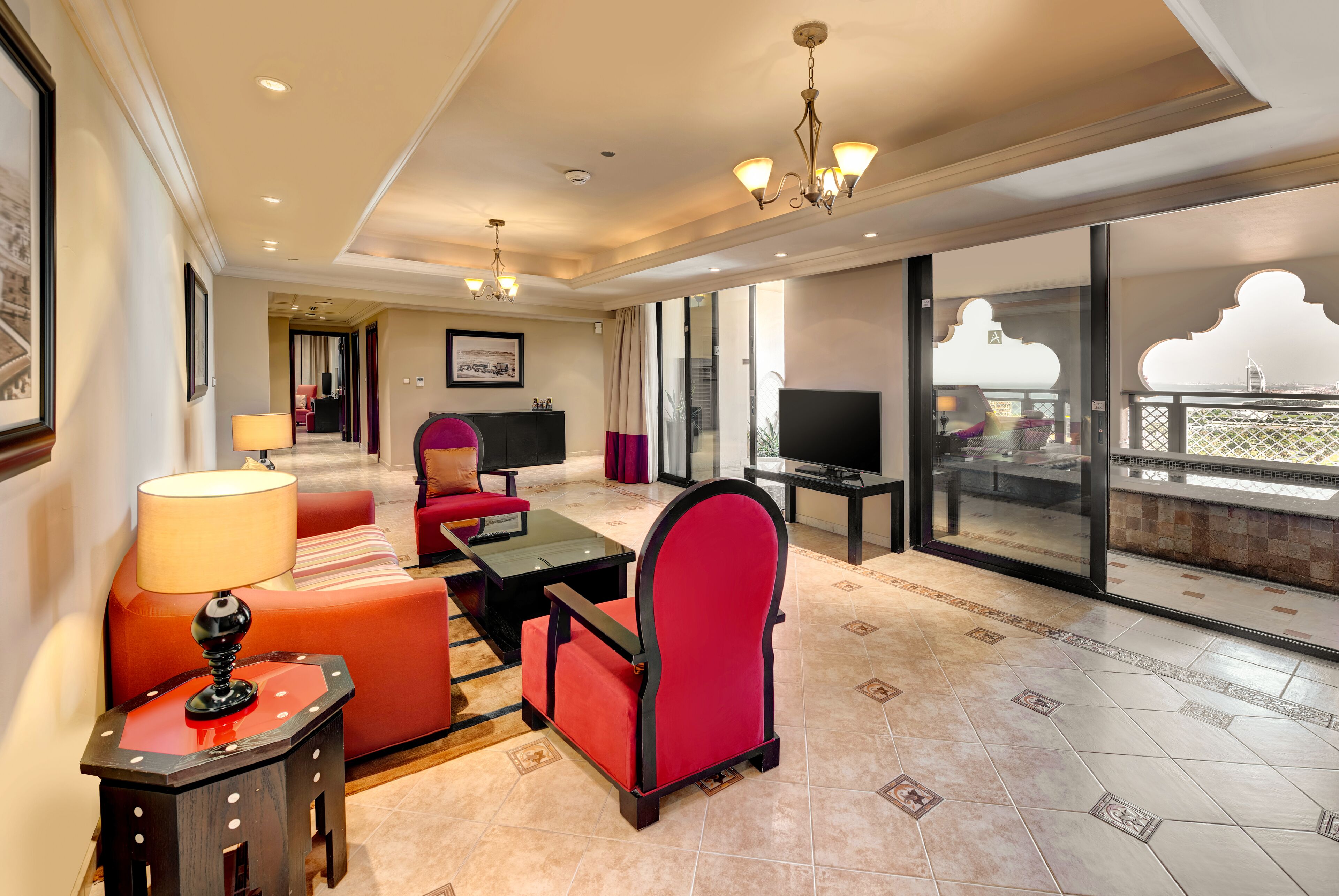 A spacious hotel room with a large balcony, a red upholstered sofa, a black armchair, and a television.