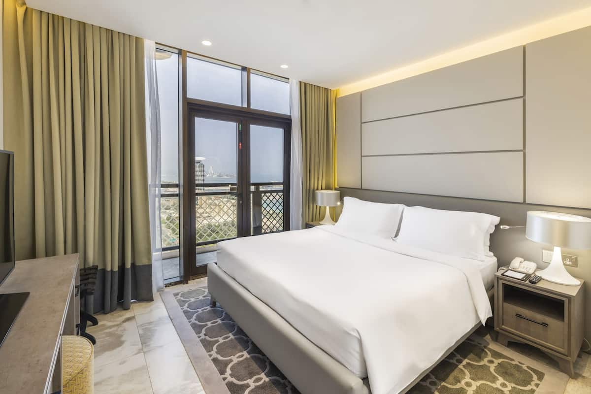 A hotel room with a large bed, a desk with a chair, a TV, a bedside table with a lamp, and a view of the city through the window.