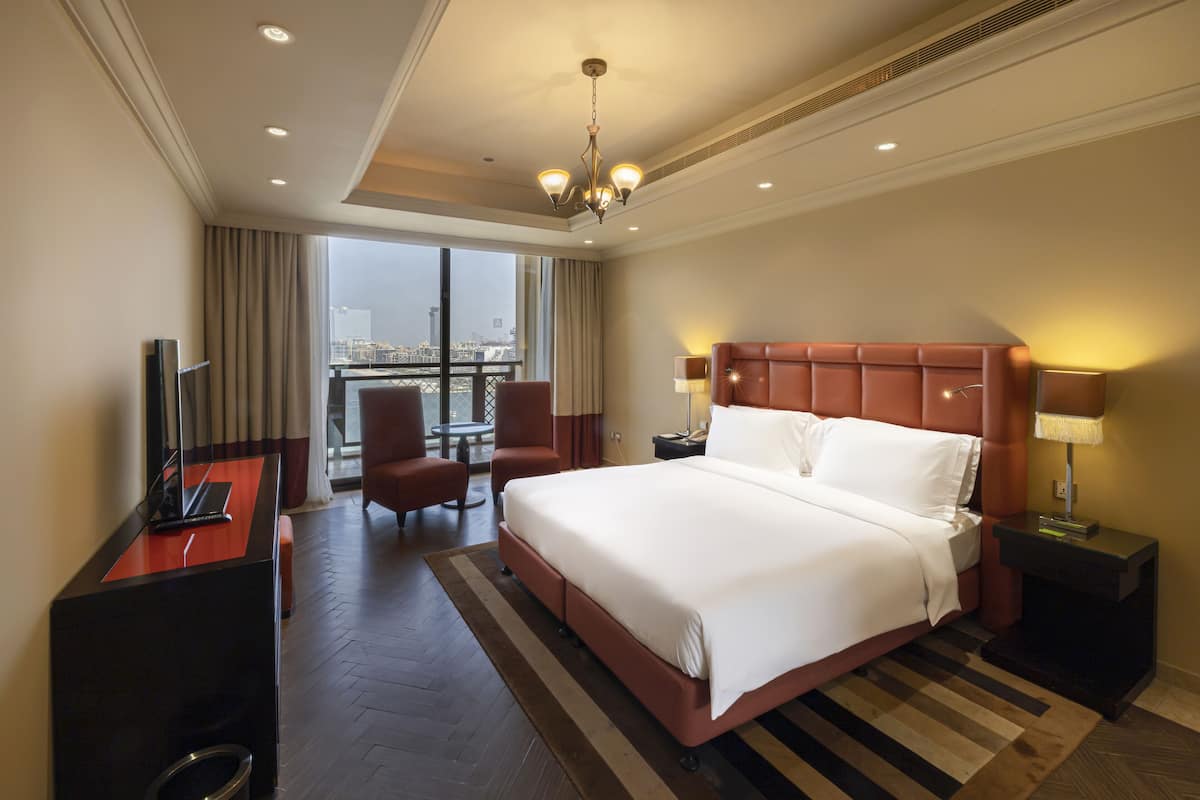A hotel room with a large bed, a desk with a laptop, a chair, a nightstand with a lamp, and a view of the city through large windows.